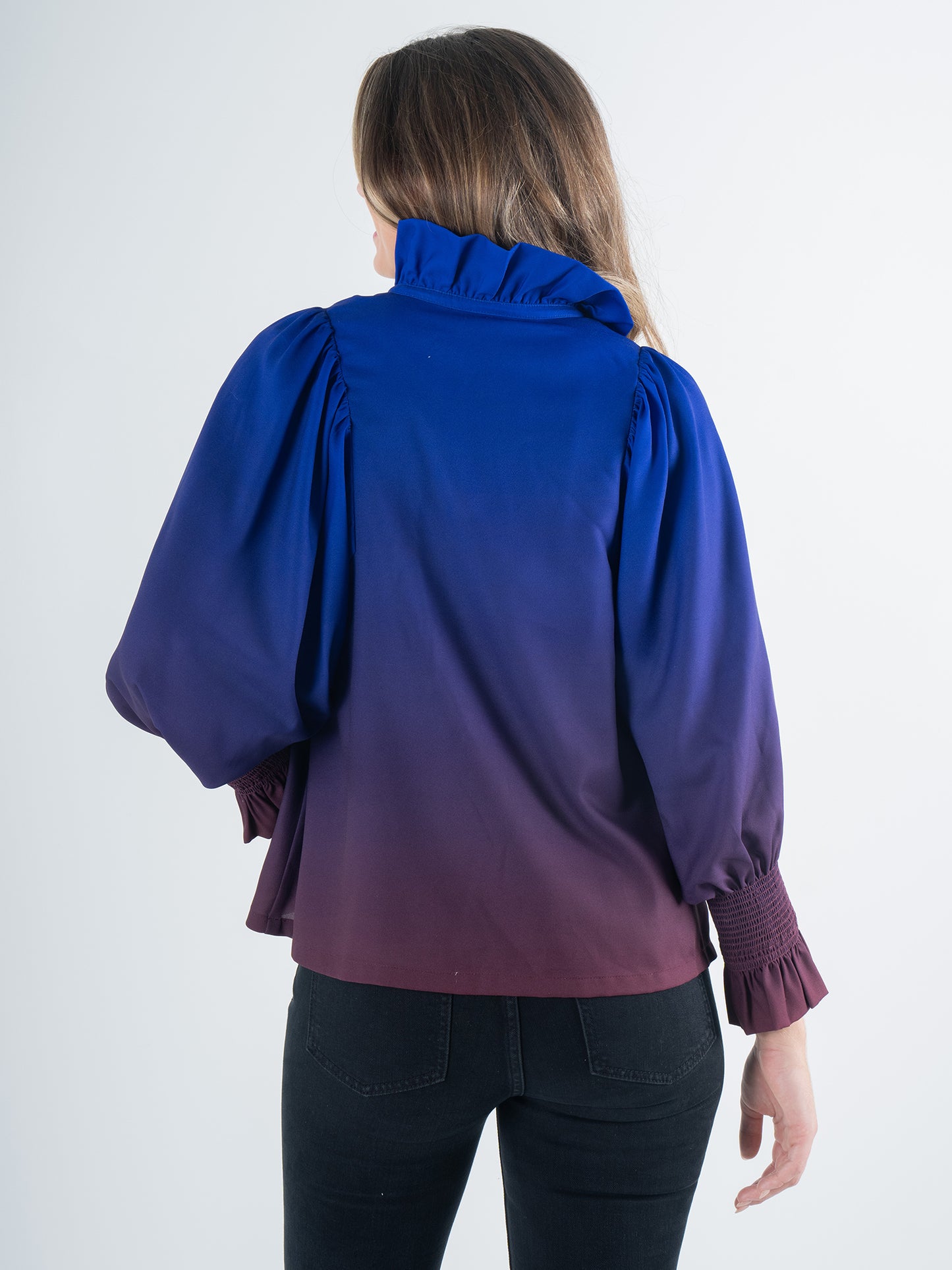 A person with long hair is wearing the Charlotte Top - Midnight Ombre, which showcases an ombre effect in purple with puff sleeves and a ruffled collar. They are turned away from the camera, paired with black pants, against a simple light background.