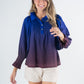 A woman smiles, wearing the Charlotte Top - Midnight Ombre with a long sleeve design in shades of blue and purple, paired with light-colored shorts. She has long hair and is standing against a plain background.