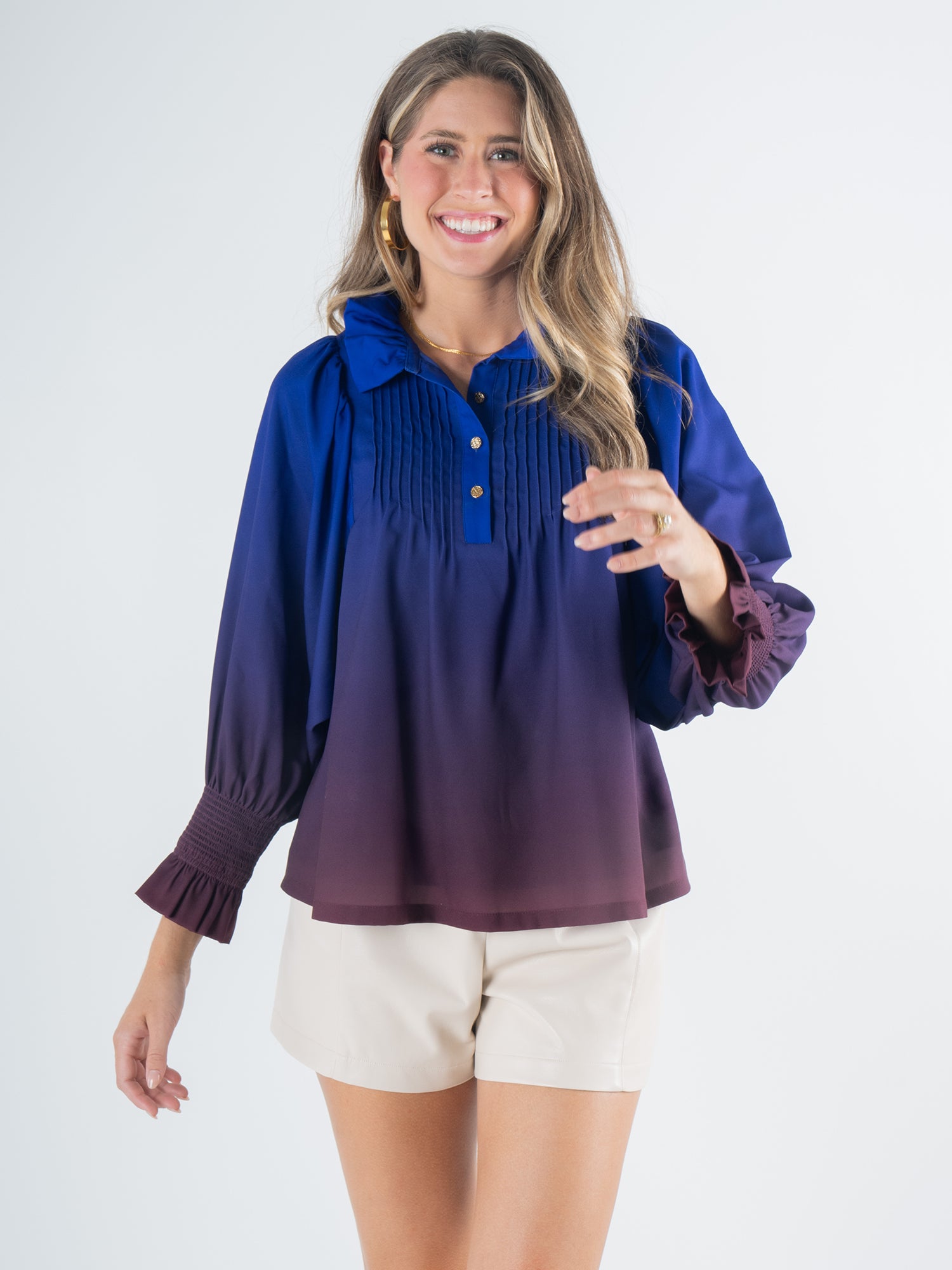 A woman smiles, wearing the Charlotte Top - Midnight Ombre with a long sleeve design in shades of blue and purple, paired with light-colored shorts. She has long hair and is standing against a plain background.