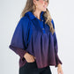 A smiling woman with long hair wears the Charlotte Top - Midnight Ombre, a blouse showcasing a blue and purple gradient with gathered details, paired with black pants. She stands against a plain white background.