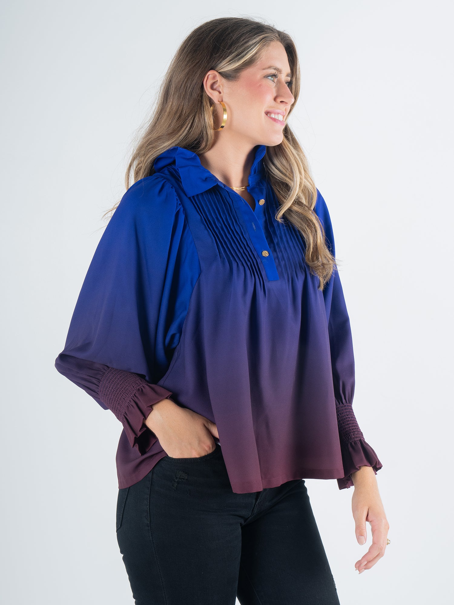 A smiling woman with long hair wears the Charlotte Top - Midnight Ombre, a blouse showcasing a blue and purple gradient with gathered details, paired with black pants. She stands against a plain white background.