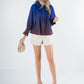 A woman in a Charlotte Top - Midnight Ombre, featuring a blue and purple gradient, pairs it with beige shorts and stands smiling. She holds a woven clutch and wears beige platform heels against a plain light-colored background.