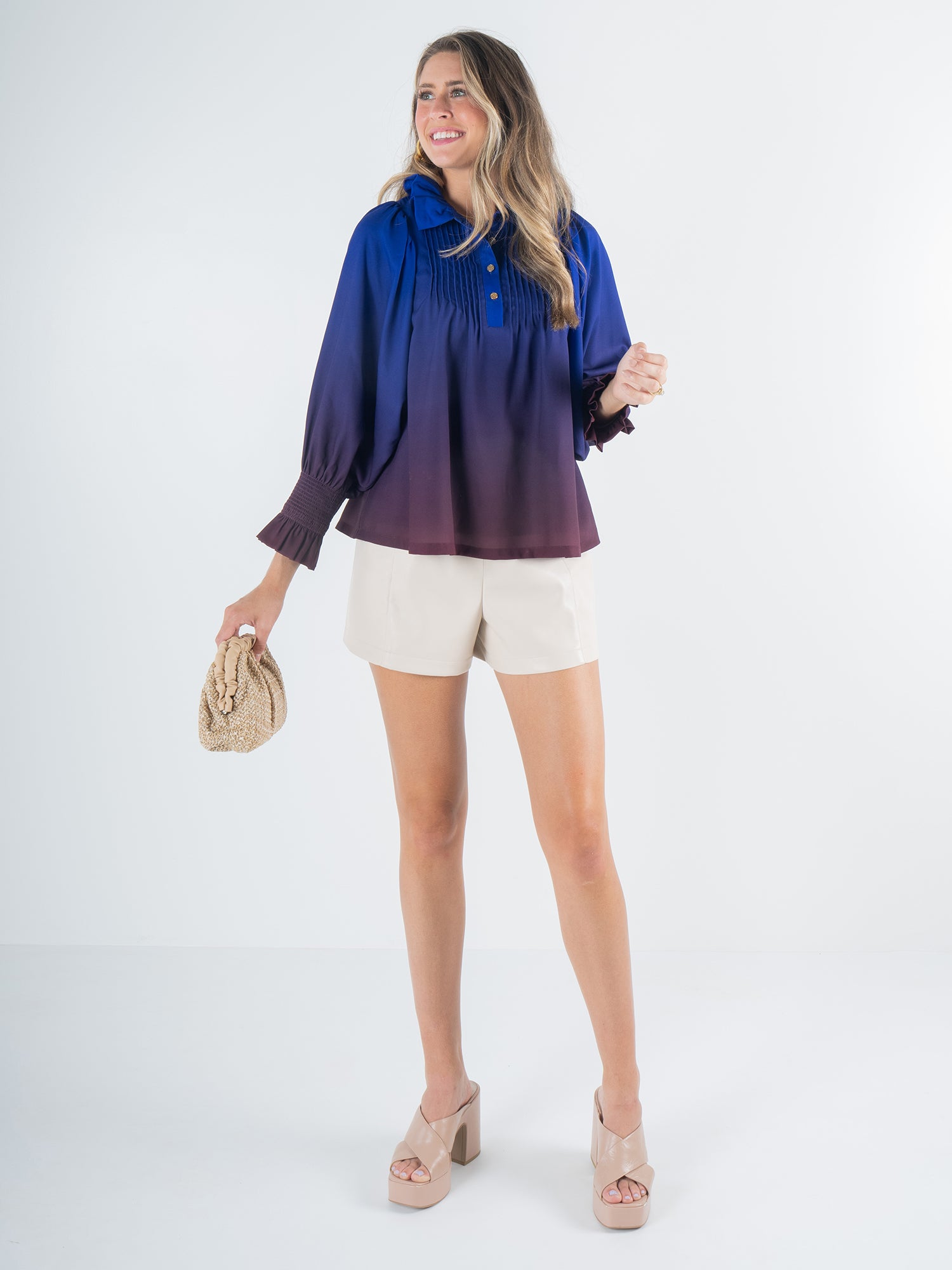 A woman in a Charlotte Top - Midnight Ombre, featuring a blue and purple gradient, pairs it with beige shorts and stands smiling. She holds a woven clutch and wears beige platform heels against a plain light-colored background.