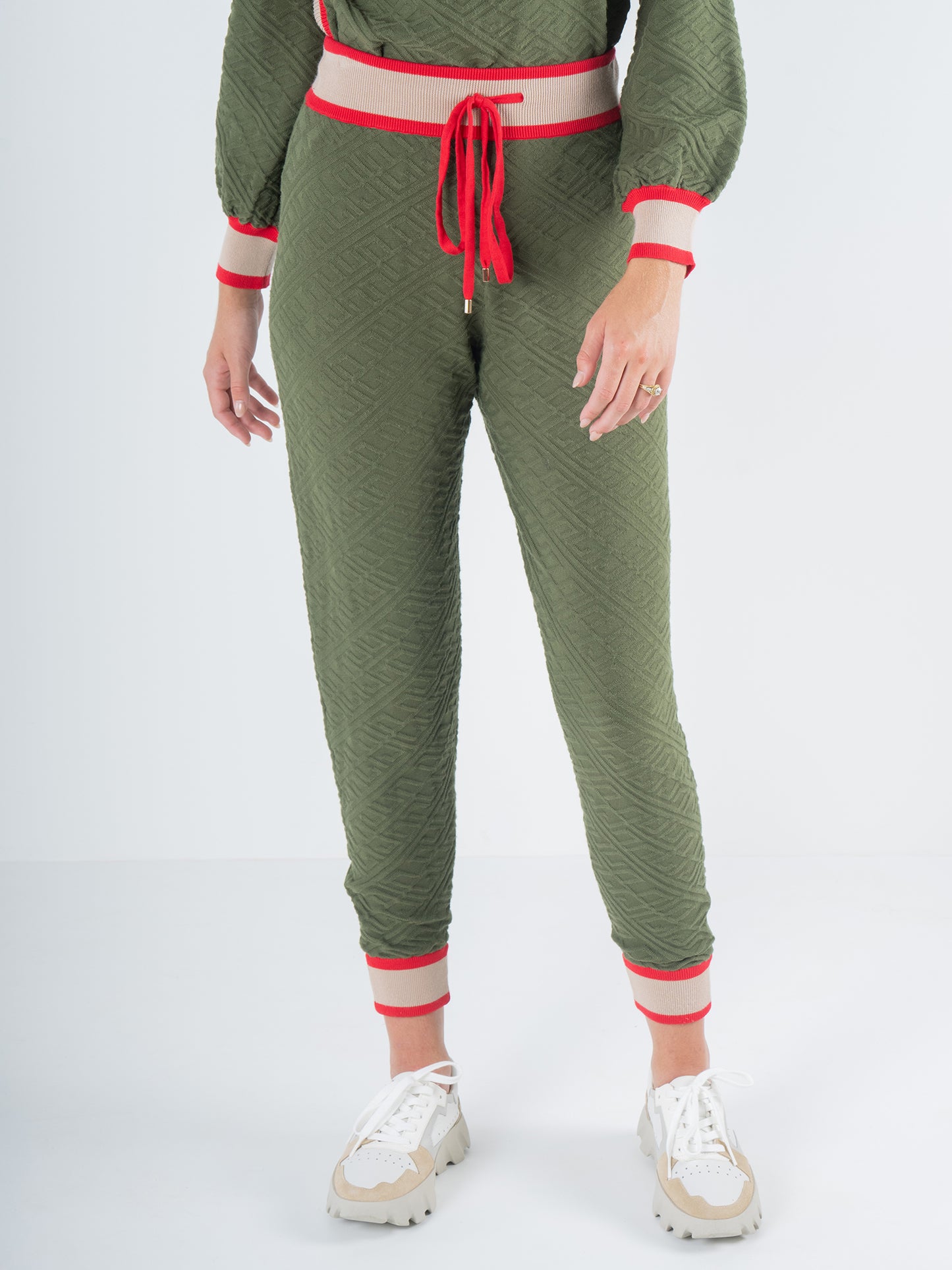 A person stands in a casual pose wearing the Downtown Jogger - Olive Night Monogram, a green tracksuit with red and white accents. The outfit includes a red waist tie and cuffs, paired with white sneakers featuring beige details. The background is a plain white setting.