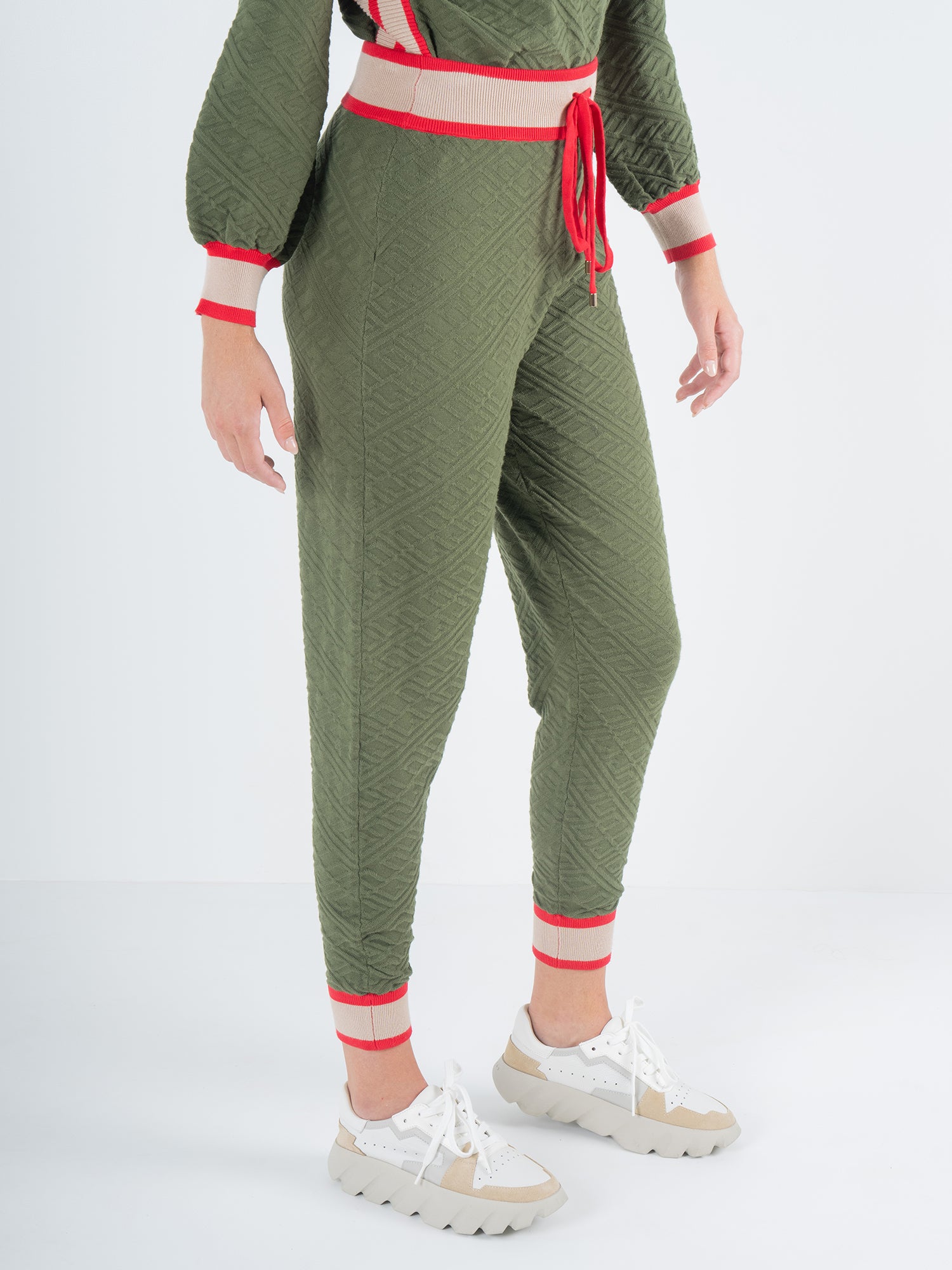 A person slips into the Downtown Jogger in Olive Night Monogram with elastic cuffs and red trim, teamed with a matching top. They finish the ensemble with chunky white sneakers featuring beige soles, ideal for stylish loungewear enthusiasts. The scene is set against a simple white studio background.