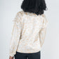 A person with curly hair stands with their back to the camera, displaying a feminine style in an Orchid Top - Neutral Freckle Spot Cheetah. This top features blouson long-sleeves and a subtle leopard print on a cream background. They pair it effortlessly with dark pants against a plain backdrop.