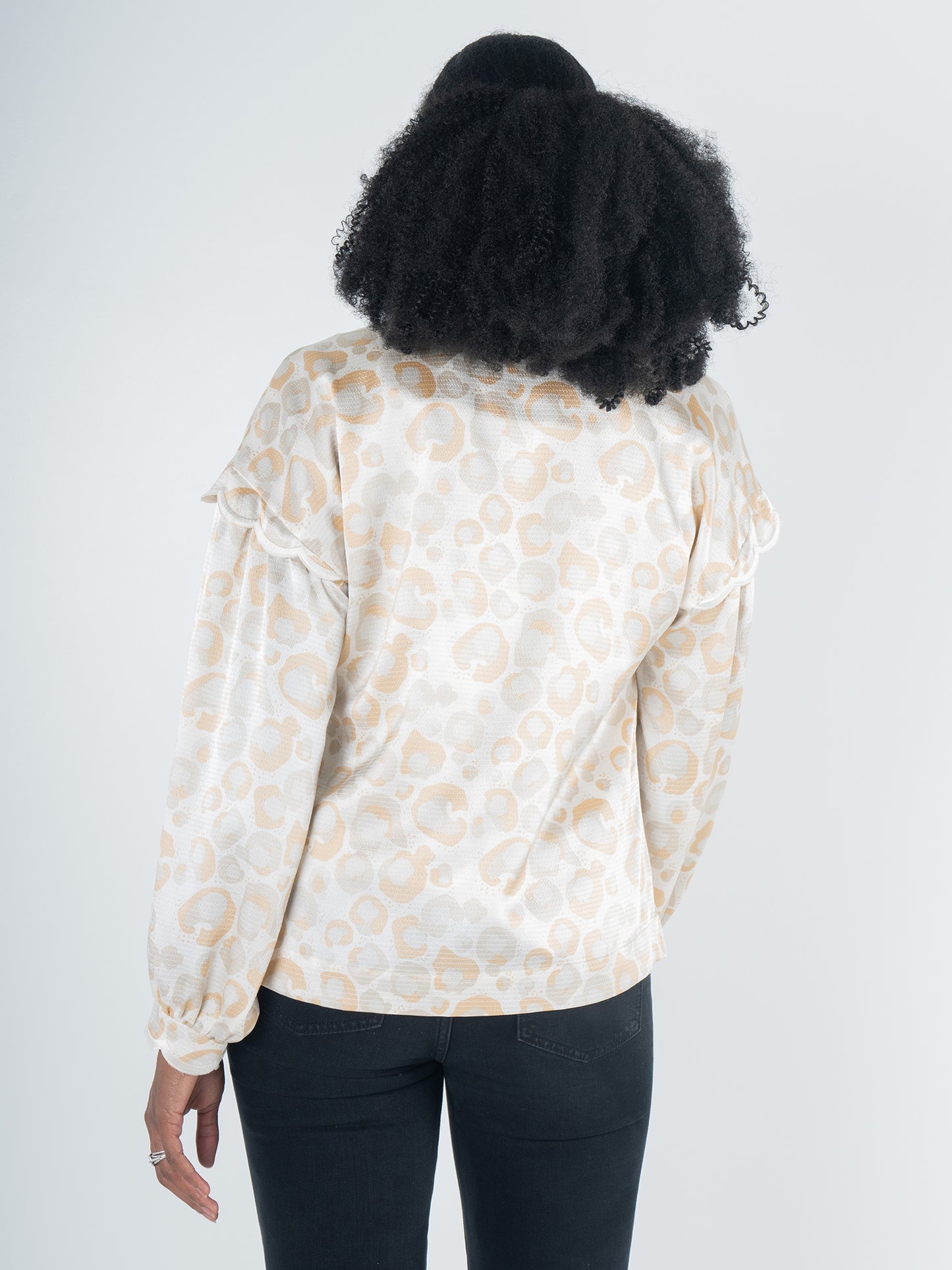 A person with curly hair stands with their back to the camera, displaying a feminine style in an Orchid Top - Neutral Freckle Spot Cheetah. This top features blouson long-sleeves and a subtle leopard print on a cream background. They pair it effortlessly with dark pants against a plain backdrop.