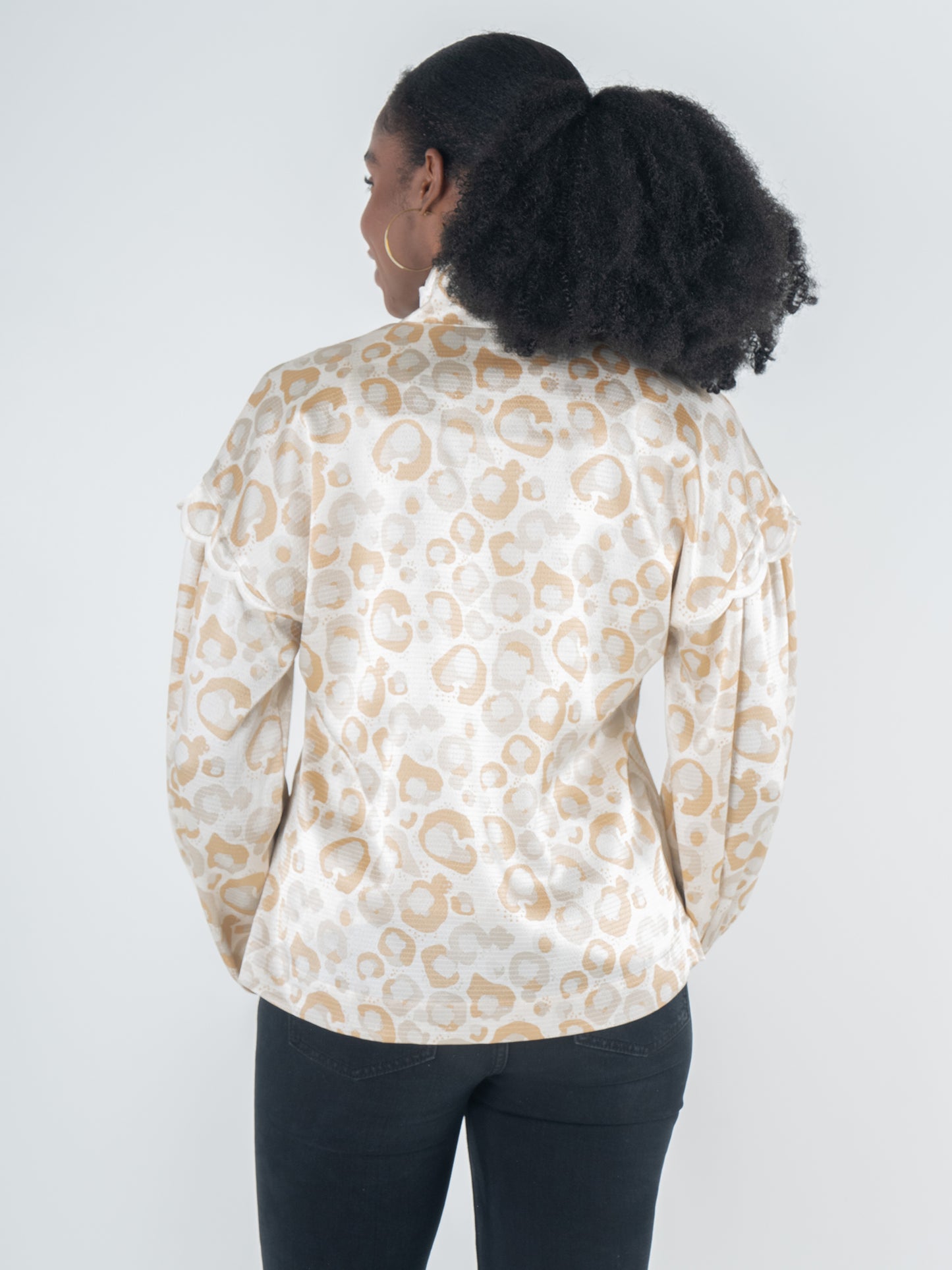 A person with dark curly hair wears the Orchid Top in a neutral freckle spot cheetah print, exuding feminine style. Their back faces the camera, highlighting the blouson long sleeves against a plain background.