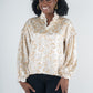 A person smiles while standing in a patterned Orchid Top - Neutral Freckle Spot Cheetah featuring blouson long-sleeves and ruffled details, paired with black pants. Their curly hair is complemented by large hoop earrings, embodying a feminine style against the plain, light-colored background.