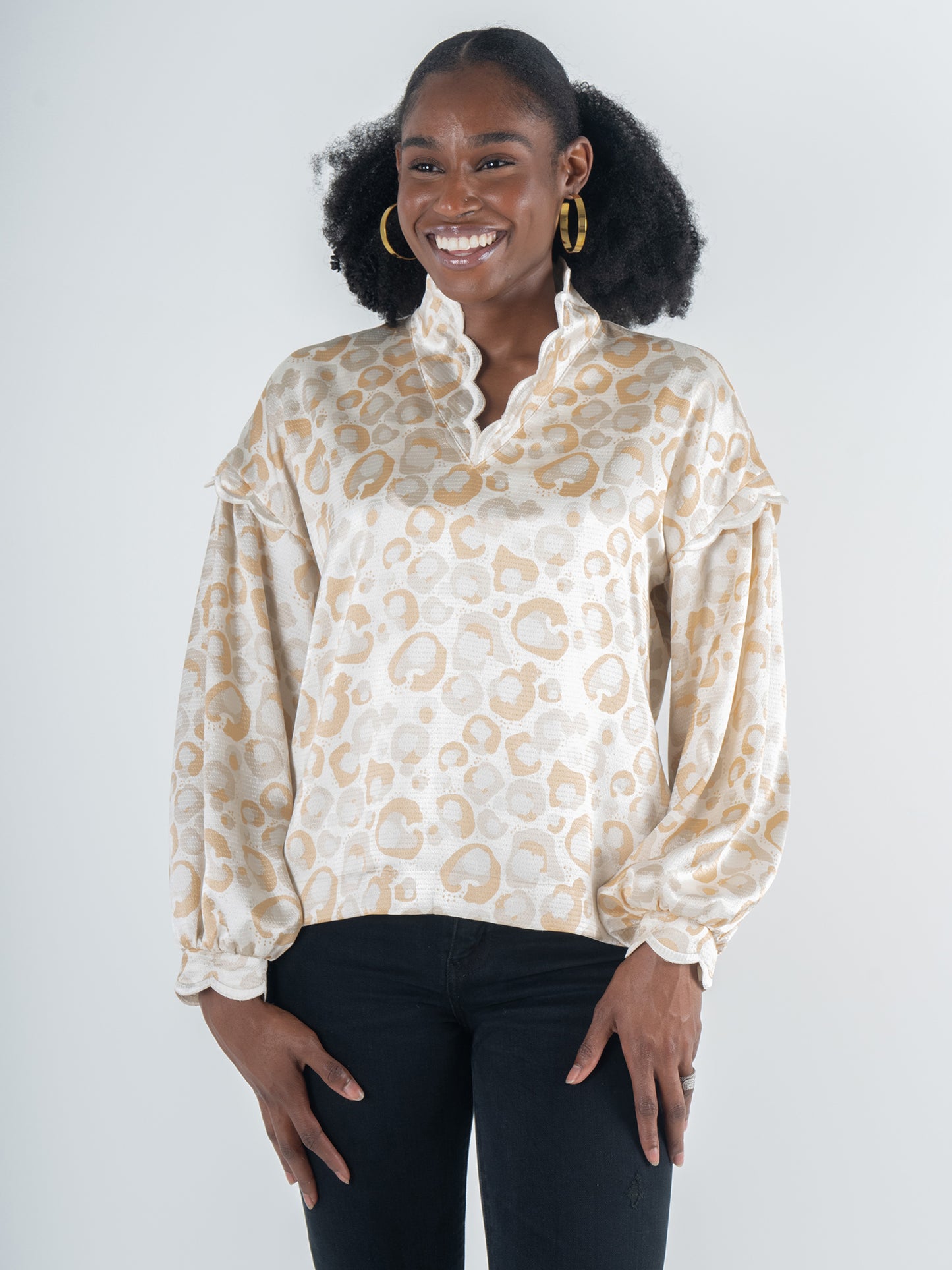 A person smiles while standing in a patterned Orchid Top - Neutral Freckle Spot Cheetah featuring blouson long-sleeves and ruffled details, paired with black pants. Their curly hair is complemented by large hoop earrings, embodying a feminine style against the plain, light-colored background.