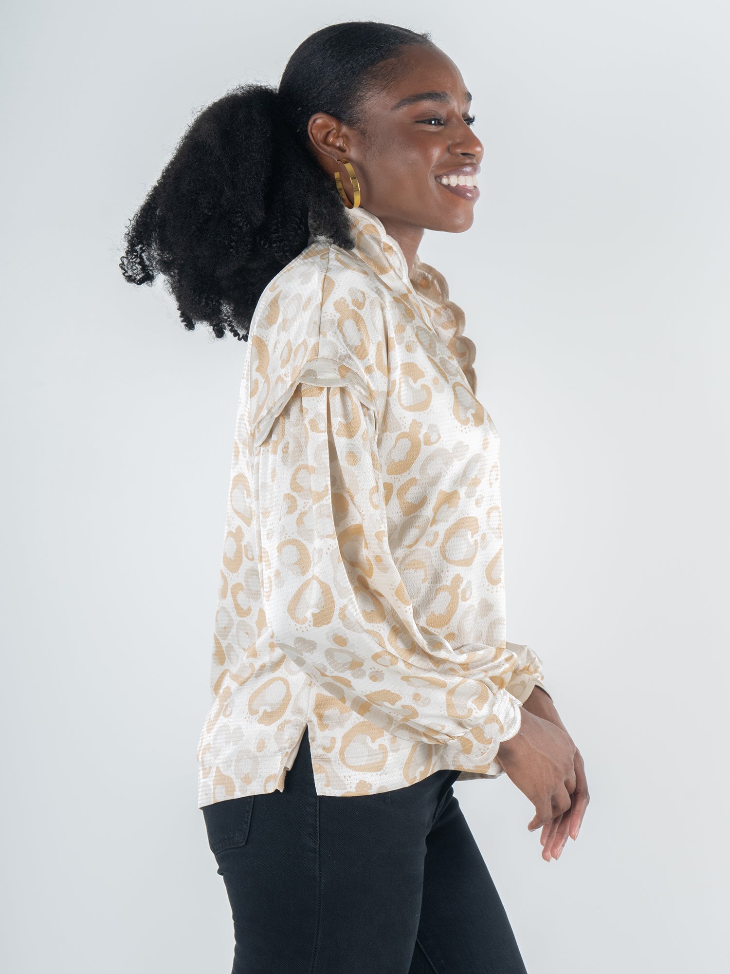 A woman with curly hair and hoop earrings is smiling in profile view. She is wearing the Orchid Top in Neutral Freckle Spot Cheetah, showcasing a feminine style, paired with black pants. The background is plain and light-colored.