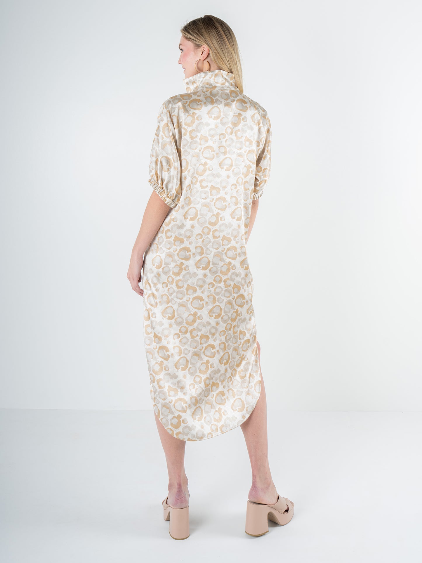 A woman with long blonde hair stands facing away, dressed in the Poppy Caftan - Neutral Freckle Spot Cheetah. Her dress features a short-sleeved design with a high collar that offers a relaxed silhouette. This look is completed with beige platform sandals in the minimalist white studio setting.