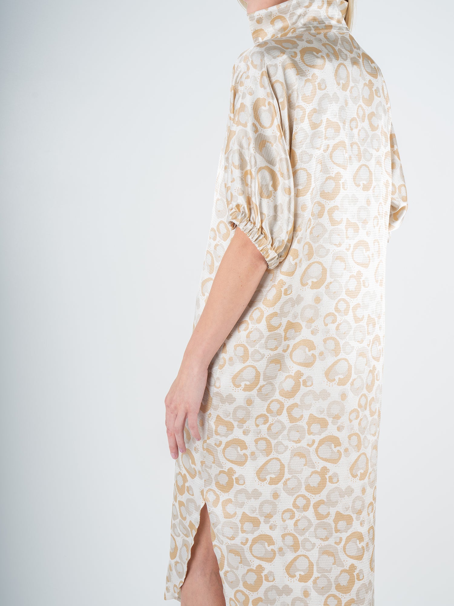 A person wearing the Poppy Caftan in Neutral Freckle Spot Cheetah, characterized by beige and white leopard print with short sleeves and a high collar, including a side slit. This relaxed silhouette stands out against the plain light gray background.