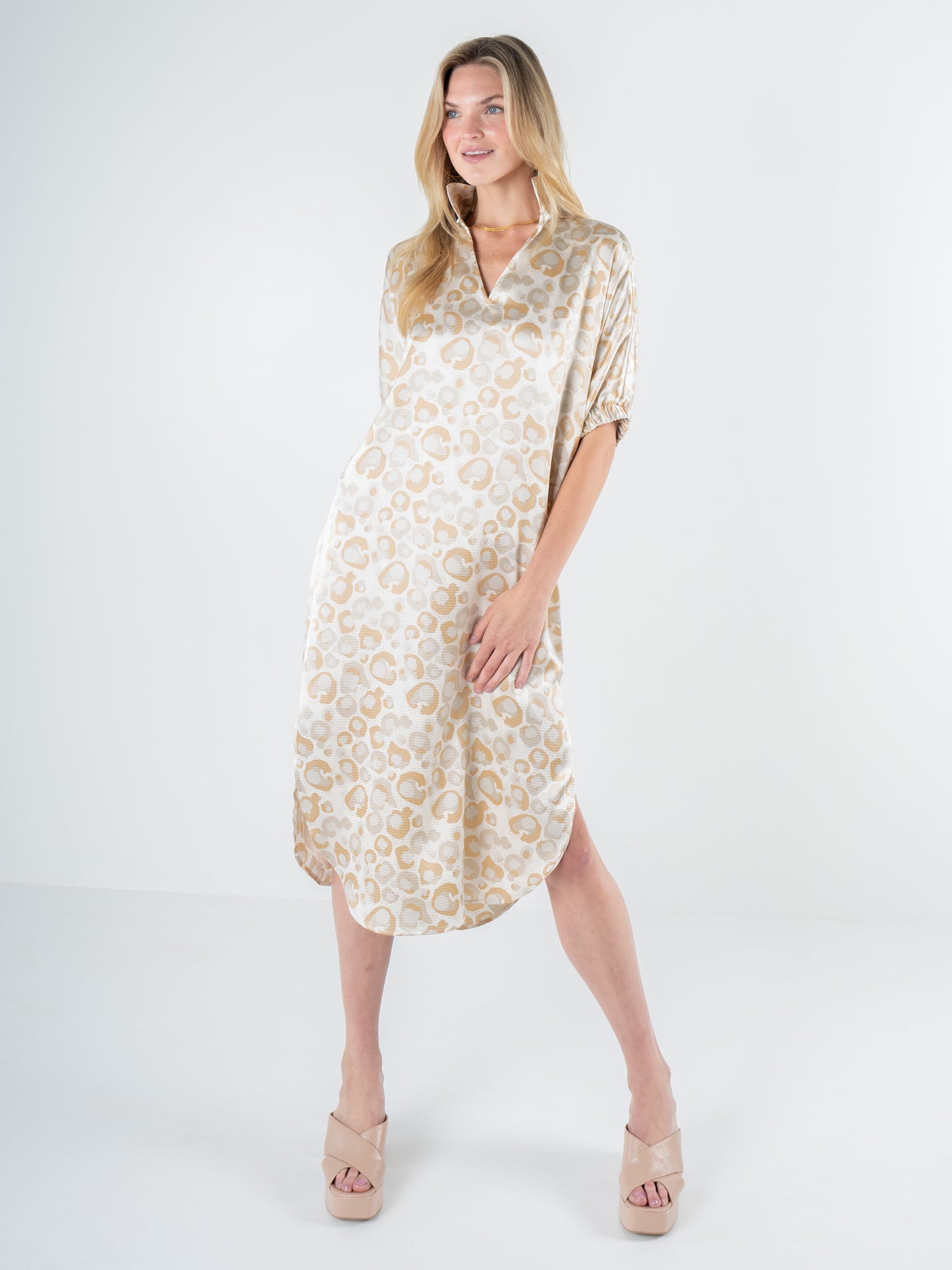 A person is posed against a plain white background, wearing the Poppy Caftan in Neutral Freckle Spot Cheetah—a loose, knee-length beige dress featuring a signature cheetah pattern. They have long hair and are wearing nude platform sandals, highlighting a relaxed silhouette that perfectly captures the style of the Poppy Caftan.