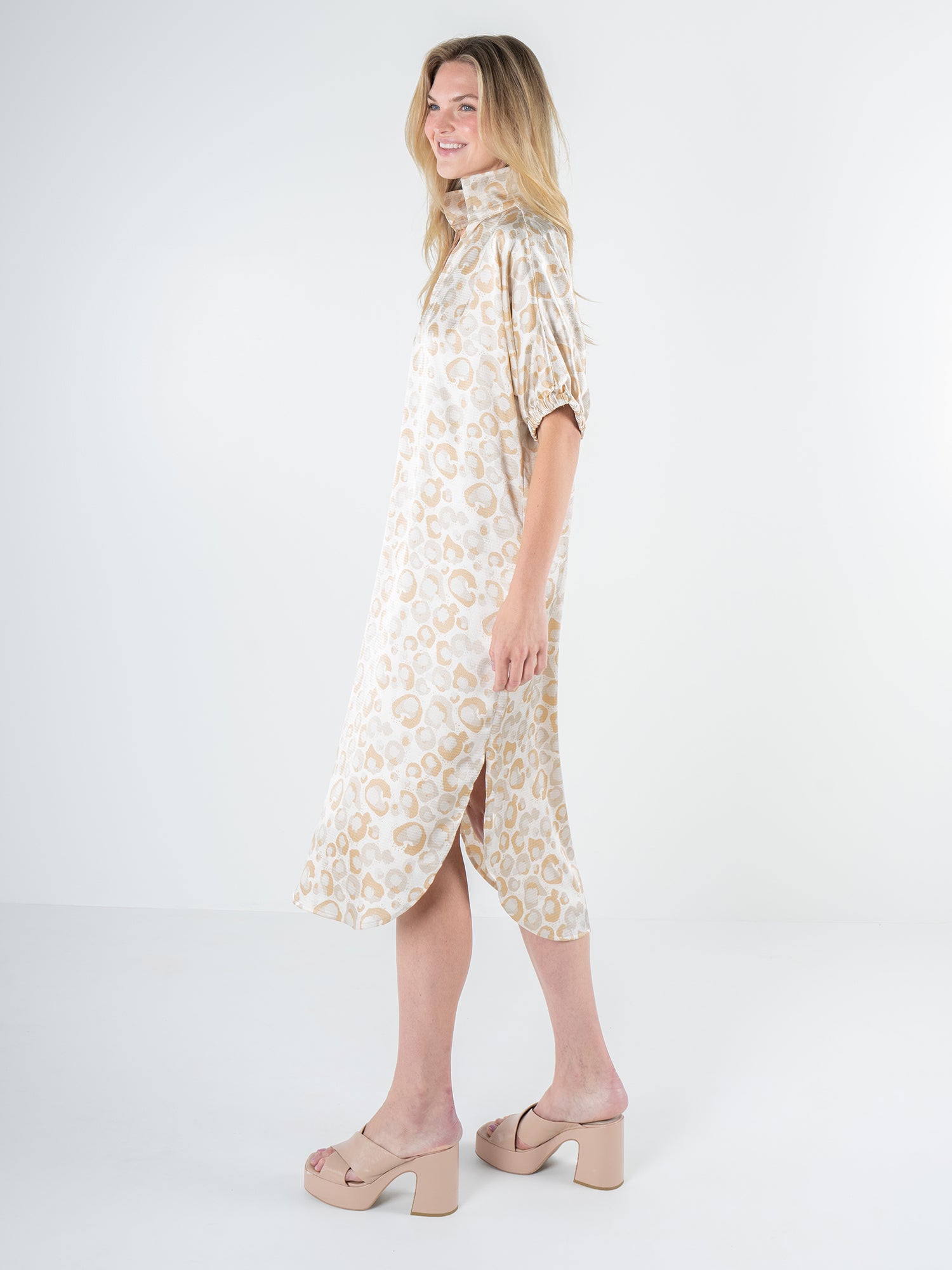 A person stands against a plain white background wearing the Poppy Caftan in Neutral Freckle Spot Cheetah. The dress features a relaxed, knee-length silhouette with a loose fit, high collar, and short sleeves. They complete the look with nude-colored platform heels, exuding effortless style.