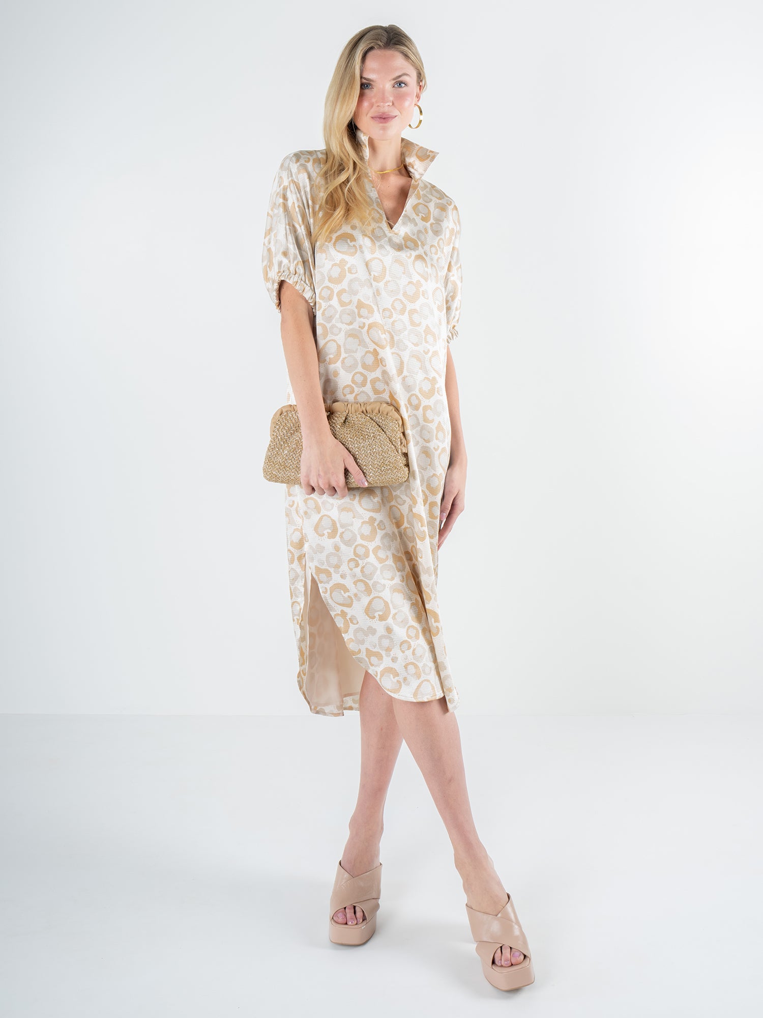 A woman stands in the Poppy Caftan - Neutral Freckle Spot Cheetah, a dress with a relaxed silhouette, holding a woven clutch. She wears matching wedge sandals and large hoop earrings, posing against a plain white background.