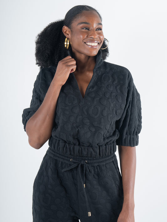 A person smiling and posing in the Poppy Crop - Jet Black Cheetah jumpsuit, featuring an embossed cheetah fabric design with a cinched waist and short sleeves. They have curly hair and are wearing hoop earrings, standing against a plain light background.