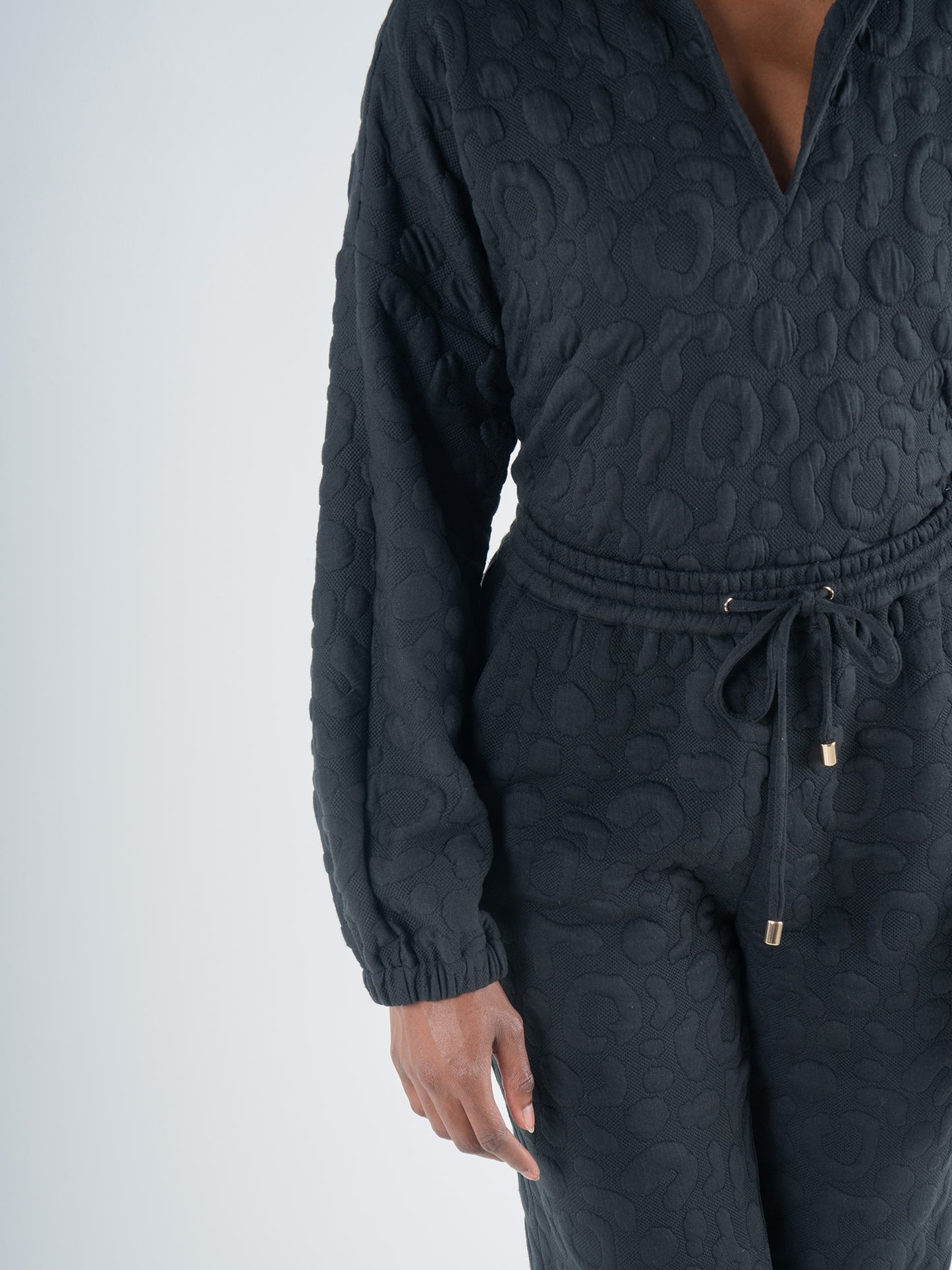 A person is wearing the Poppy Pop-Over in Jet Black Cheetah, a textured long-sleeve jumpsuit made from embossed cheetah fabric featuring a distinct pattern and drawstring waist, as they pose against a simple light background. The image highlights the upper half of their body and right arm.