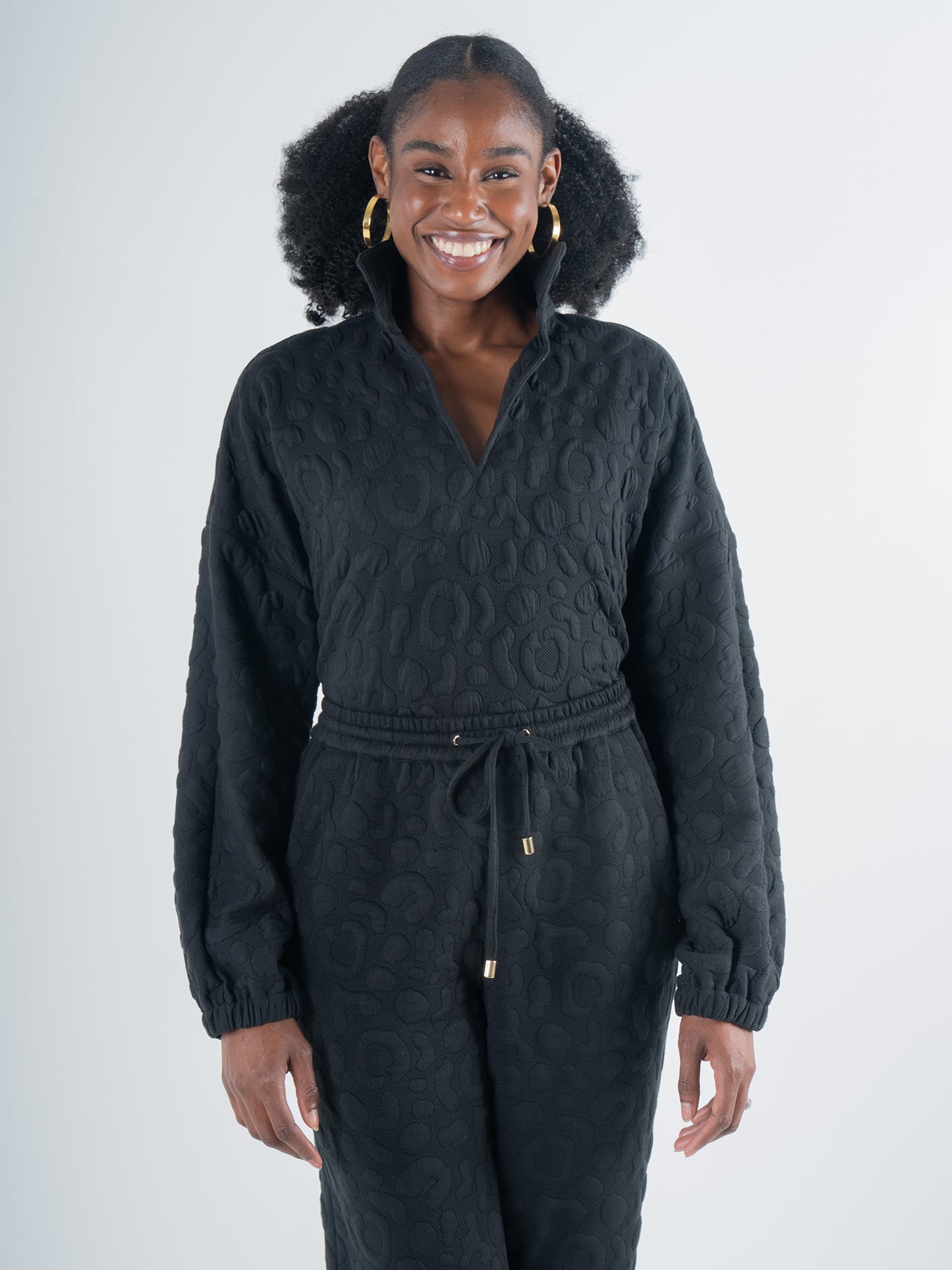 A person smiling, dressed in the Poppy Pop-Over in Jet Black Cheetah, which features a high collar and embossed cheetah fabric with matching pants. The understated pattern enhances their natural curls and gold earrings, set against a plain, soft gray background that adds elegance to the ensemble.