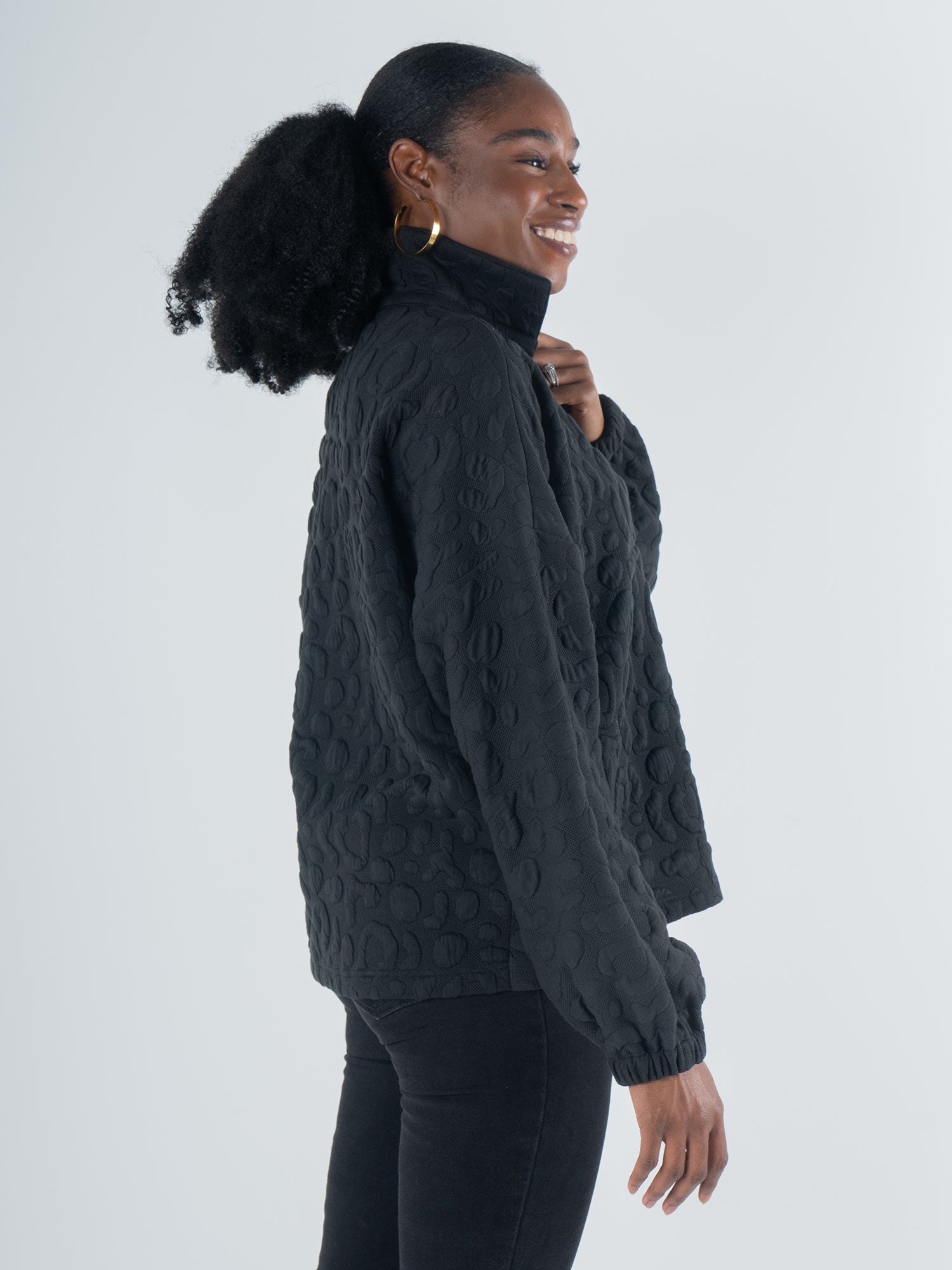 A person with long, curly hair tied back is smiling and standing in profile. They are wearing the Poppy Pop-Over in Jet Black Cheetah and hoop earrings, set against a light gray background.