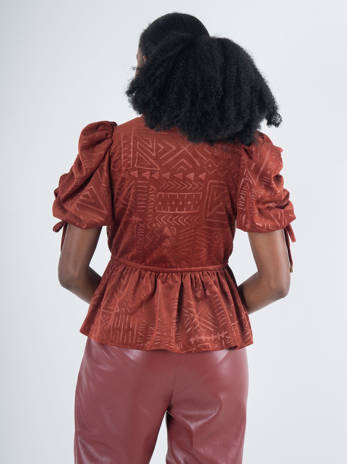 A person with curly hair faces away, exuding bohemian charm in a Posey Top - Ancient Reverie, featuring puffed sleeves and a geometric pattern in rust color, paired with matching leather pants. The background is a soft, pale hue.