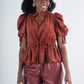 A woman exudes bohemian charm in a rust-colored Posey Top - Ancient Reverie featuring puff sleeves and a gathered waist, paired with matching wide-leg pants. Her curly hair is styled in two puffs, and she accessorizes with large hoop earrings against a plain light gray backdrop.