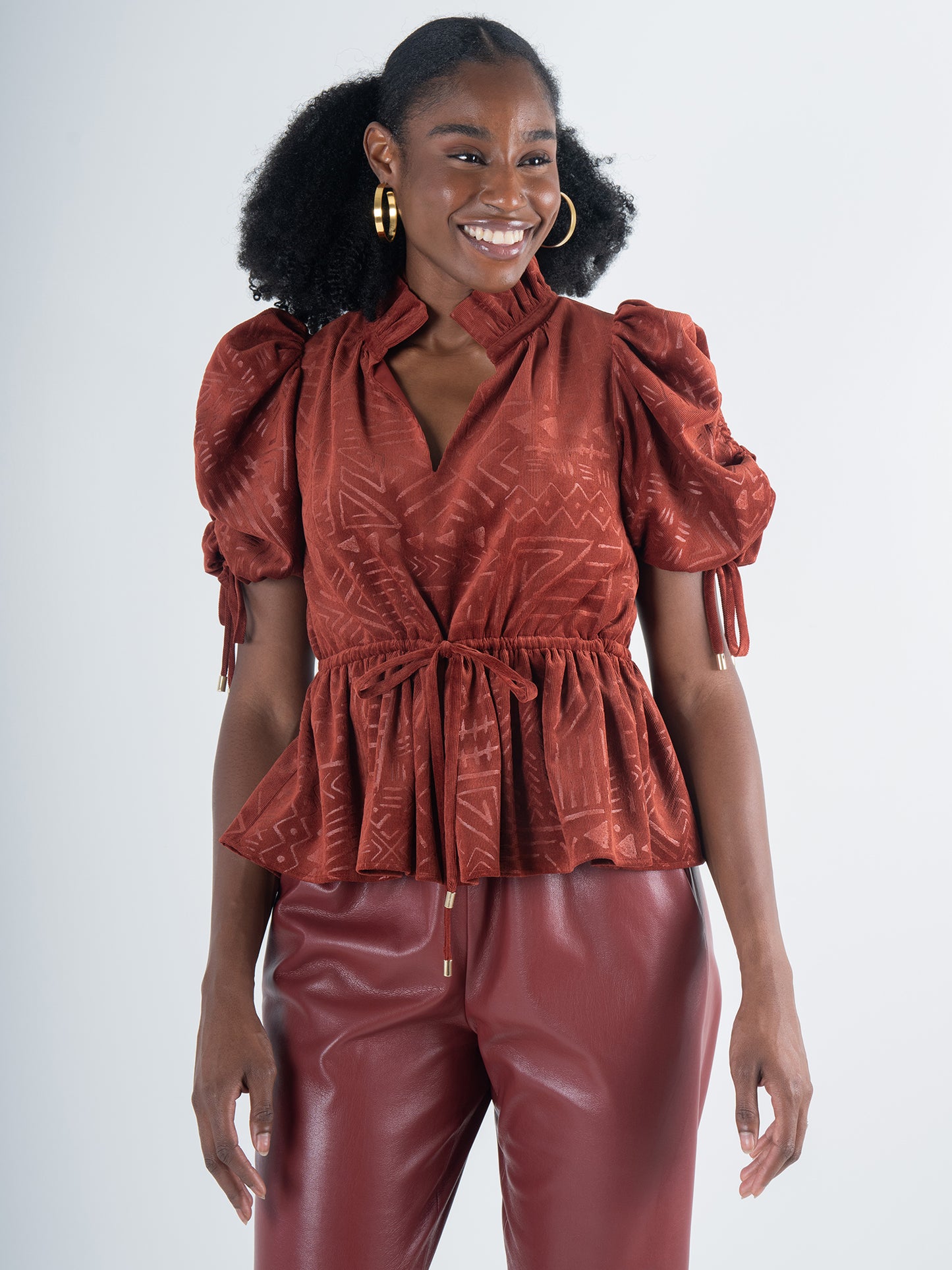 A woman exudes bohemian charm in a rust-colored Posey Top - Ancient Reverie featuring puff sleeves and a gathered waist, paired with matching wide-leg pants. Her curly hair is styled in two puffs, and she accessorizes with large hoop earrings against a plain light gray backdrop.