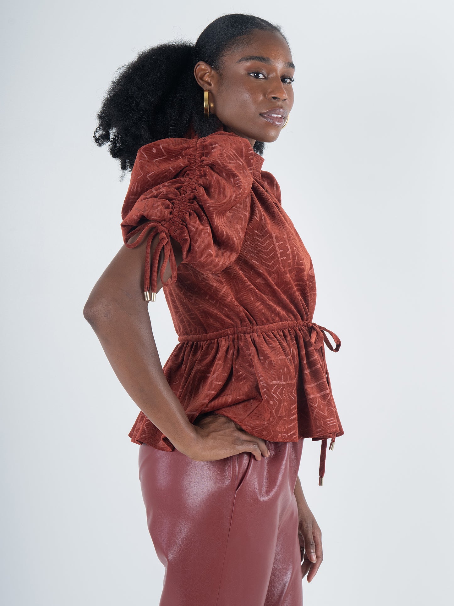 A woman stands in profile wearing the Posey Top - Ancient Reverie, showcasing puffed sleeves and a drawstring waist, paired with matching rust-colored pants. Her hair is styled in a natural puff that enhances her bohemian charm, complemented by gold hoop earrings against a plain background.