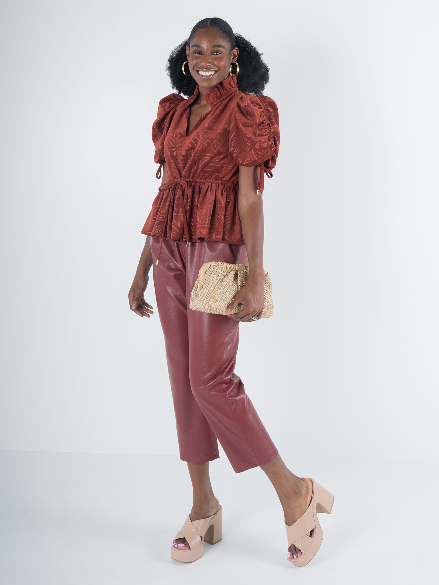 A woman wearing an Ancient Reverie Posey Top in rust and matching leather pants holds a woven clutch. Exuding bohemian charm, she stands in a light-colored space, smiling, with her black curly hair tied back. She completes the look with heeled sandals and gold hoop earrings.