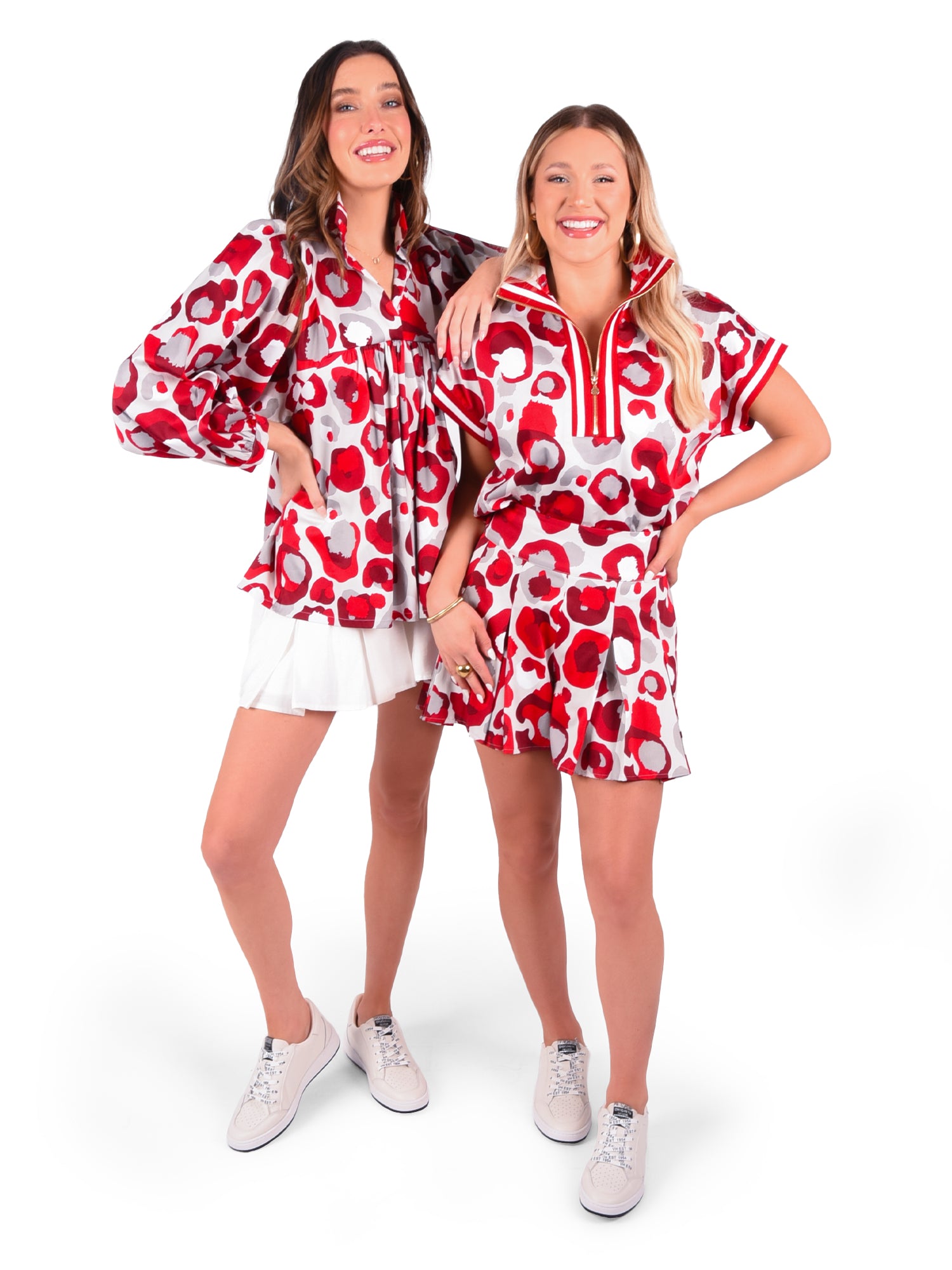 Two women are smiling and standing side by side, wearing matching red, white, and pink patterned outfits. One is dressed in a blouse with dolman sleeves paired with a white skirt, while the other sports the short-sleeved Poppy Pullover in Burgundy Collegiate Cheetah with shorts. Both complete their looks with sleek white sneakers.