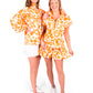 Two women are smiling and posing together in matching outfits featuring the Poppy Pullover in Orange Collegiate Cheetah pattern. One wears a long-sleeved top with a 3/4 zip paired with a skirt, while the other sports a short-sleeved dress. Both complement their look with white sneakers against a pristine white background.