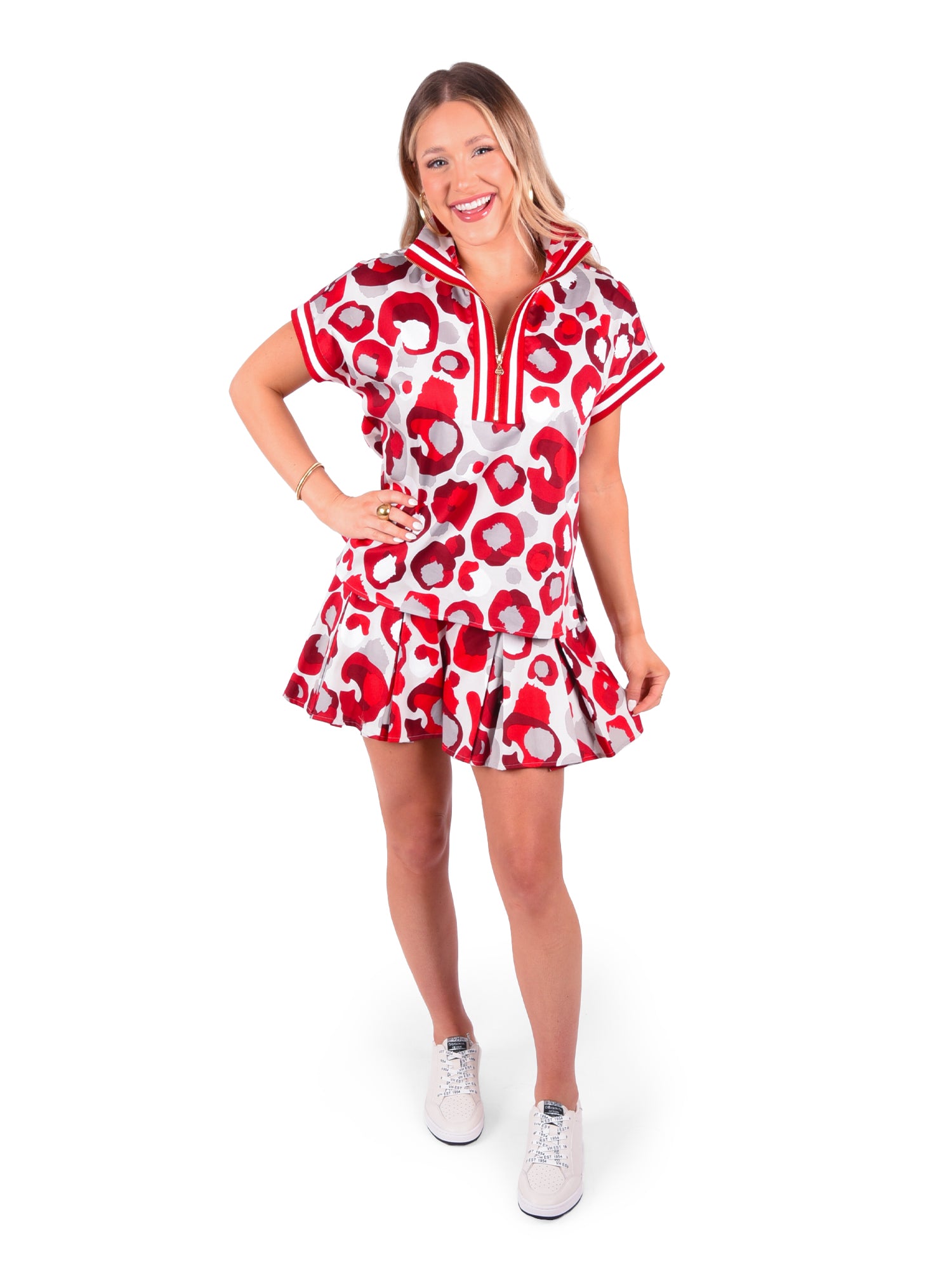 A woman is smiling and posing in a chic dress with short sleeves, displaying a striking burgundy collegiate cheetah pattern. The dress boasts a flared skirt and pairs effortlessly with white sneakers, perfectly capturing the charm of the Poppy Pullover against the pristine, white backdrop.