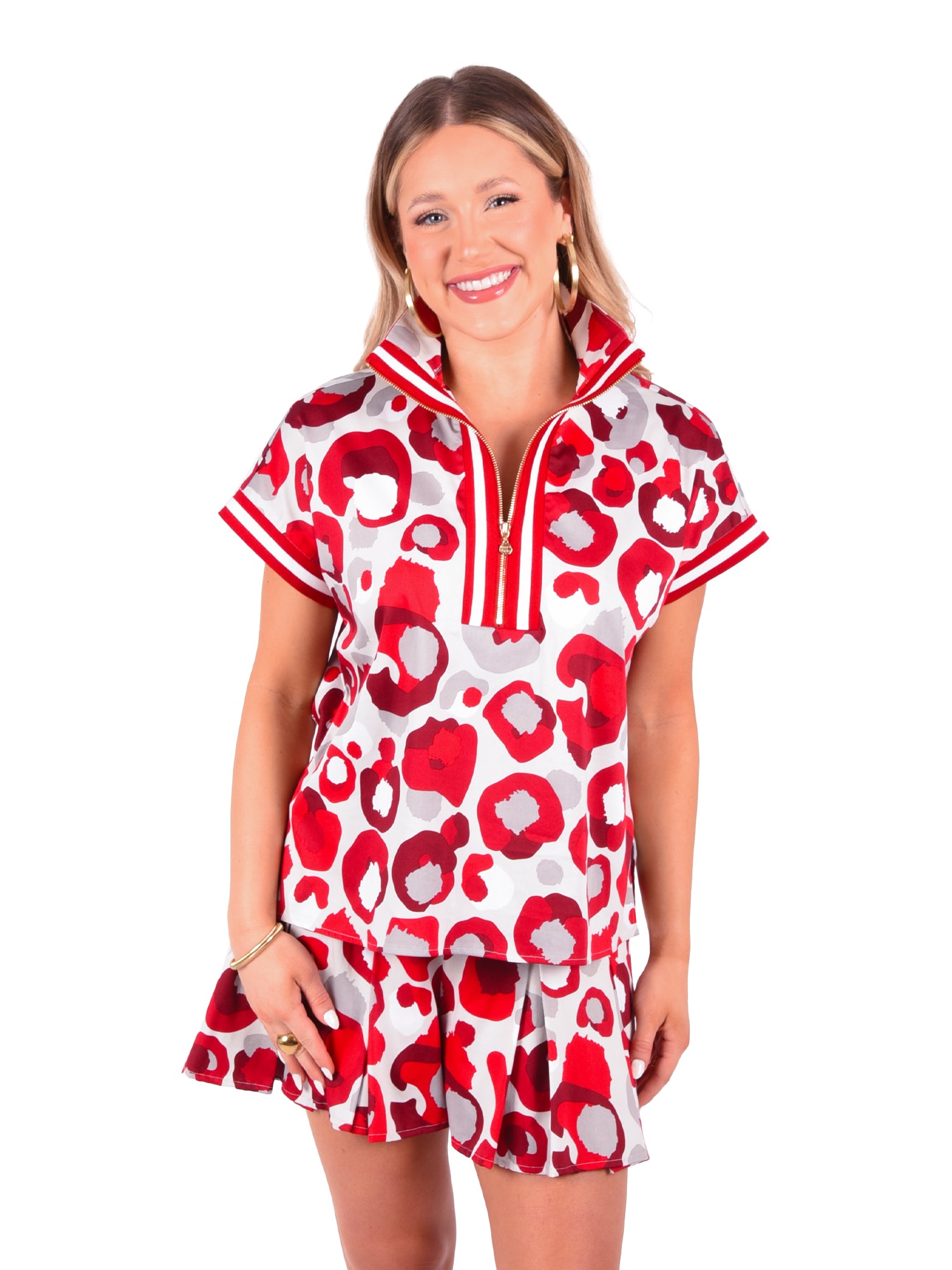 A smiling woman showcases a vibrant burgundy and white patterned outfit, featuring a Poppy Pullover in the Collegiate Cheetah design with a 3/4 zip top and matching skirt. Her long hair cascades down, complemented by gold hoop earrings, as she stands against a crisp white background.