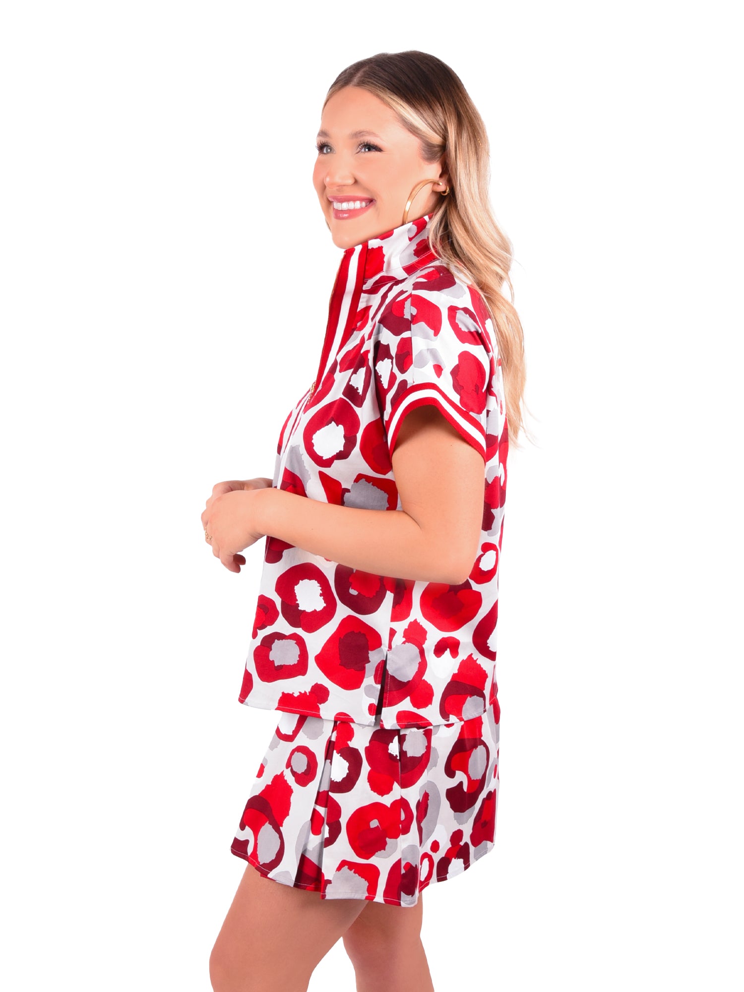 A person smiling while wearing a matching set adorned with the burgundy collegiate cheetah pattern. The Poppy Pullover showcases a short-sleeved top with a high collar, perfectly paired with a skirt. The plain white background accentuates the striking ensemble.