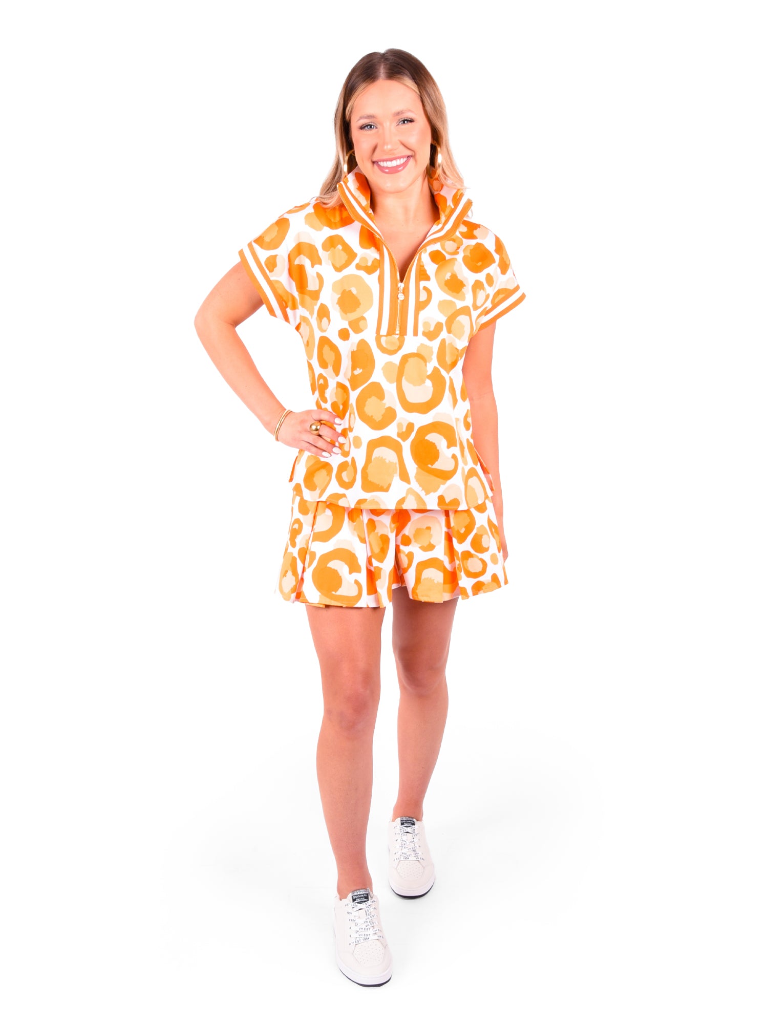 A person smiling stands with one hand on their hip, wearing the sleek and stylish Poppy Pullover in Orange Collegiate Cheetah. The short-sleeved, zip-up dress showcases an eye-catching orange and white abstract pattern, perfectly complemented by their choice of white sneakers. The simple white background accentuates the vibrancy of their attire.