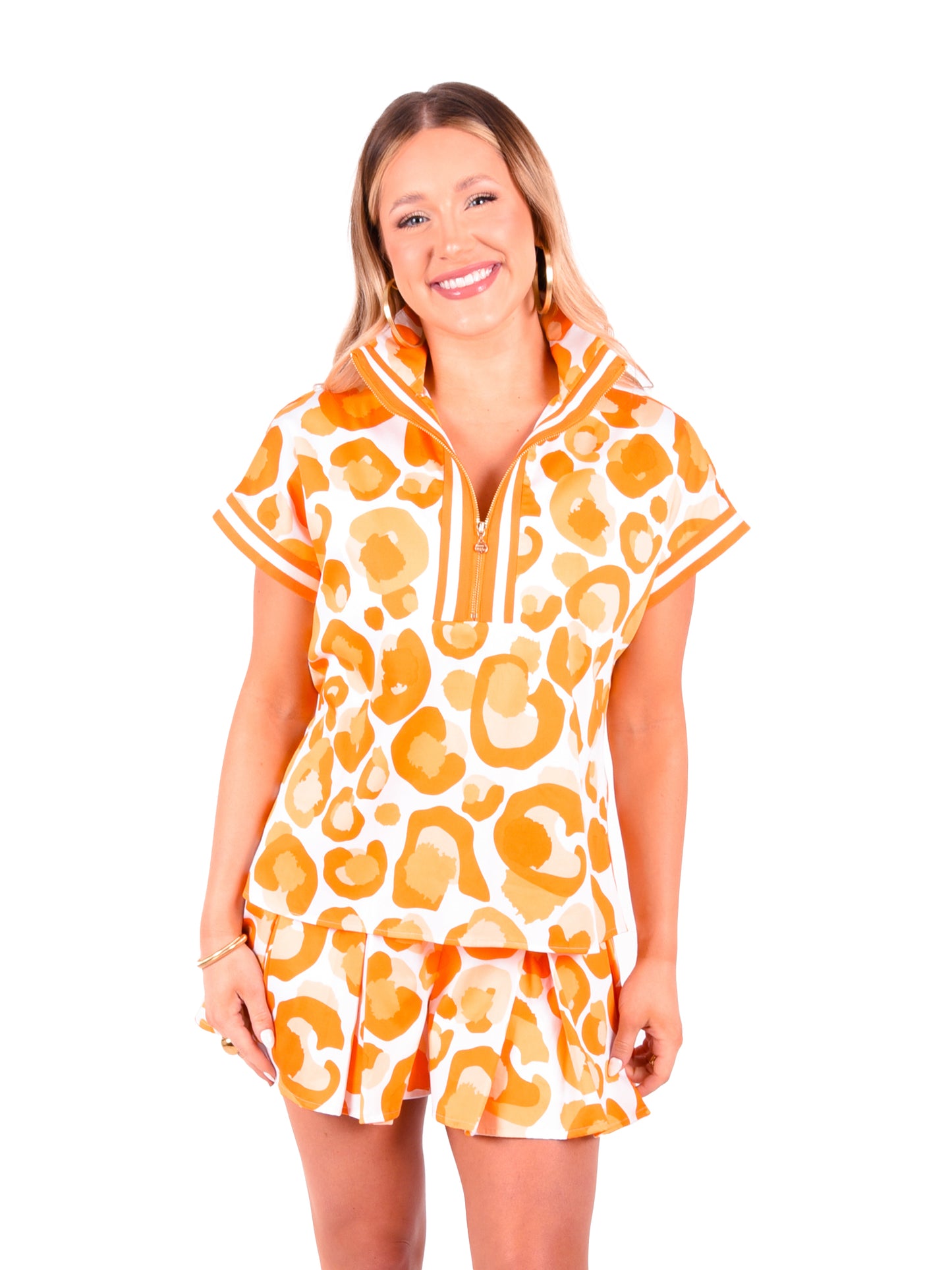 A person with long hair is smiling while wearing a skirt and the Poppy Pullover - Orange Collegiate Cheetah, which features an orange and white abstract pattern along with a stylish 3/4 zip neckline. They are posing against a plain white background.