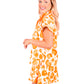 A woman with long blonde hair smiles and stands sideways, showcasing the Poppy Pullover in Orange Collegiate Cheetah with dolman sleeves, paired with a matching skirt. Set against a plain white background, she embodies an effortlessly stylish look.