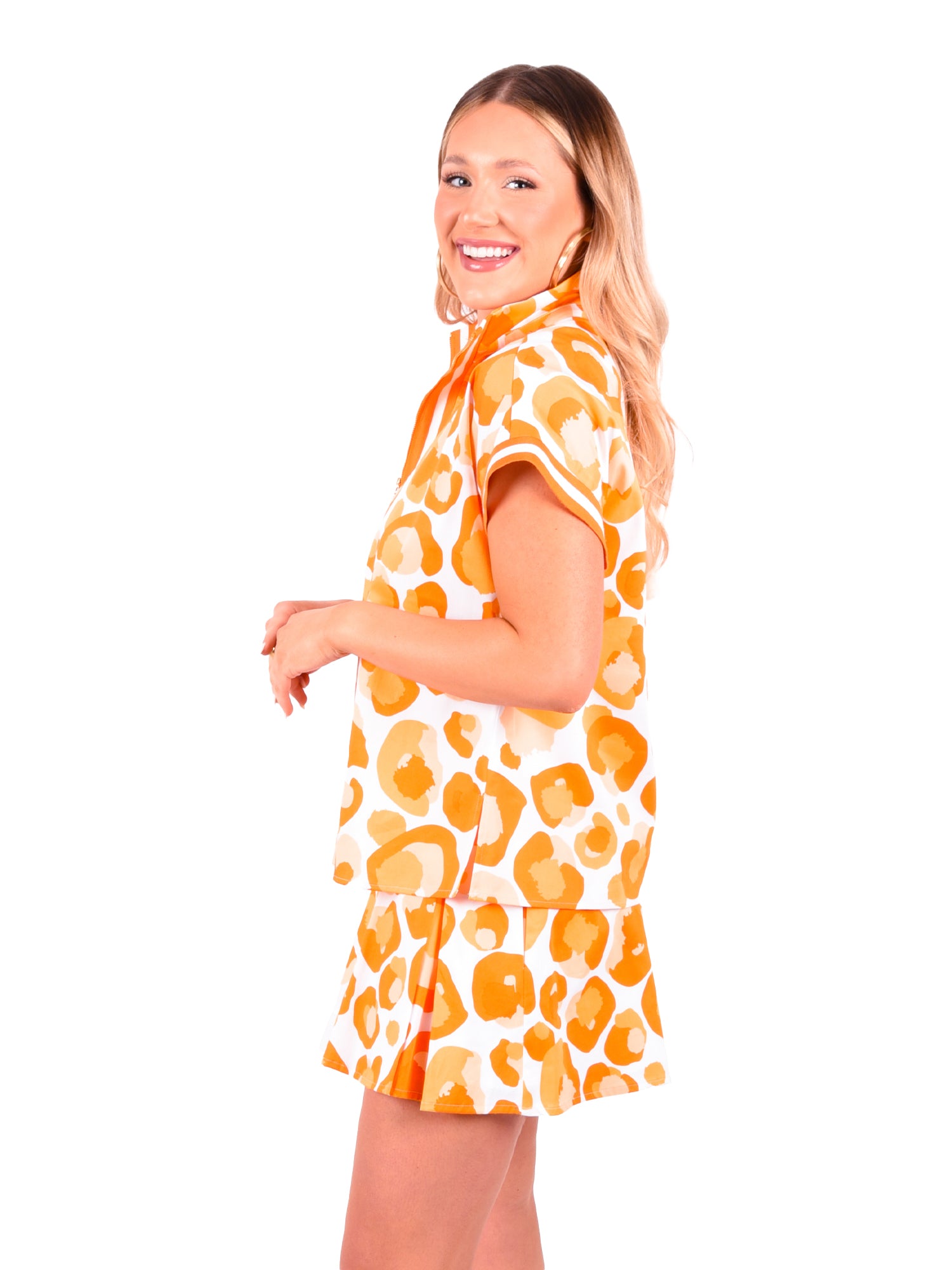 A woman with long blonde hair smiles and stands sideways, showcasing the Poppy Pullover in Orange Collegiate Cheetah with dolman sleeves, paired with a matching skirt. Set against a plain white background, she embodies an effortlessly stylish look.