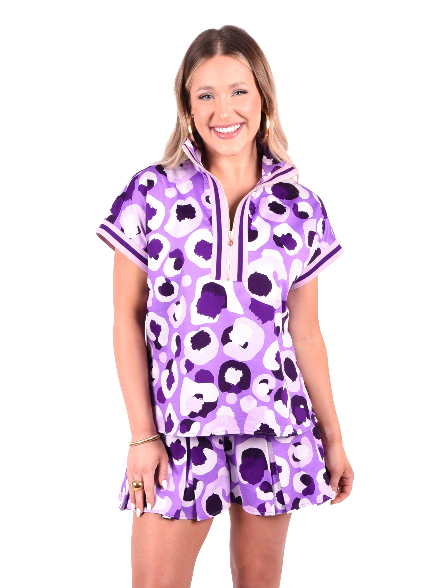 A person smiling, wearing the Poppy Pullover in Purple Collegiate Cheetah with dolman sleeves and a short skirt. The top showcases a zippered neckline, set against a plain white background.