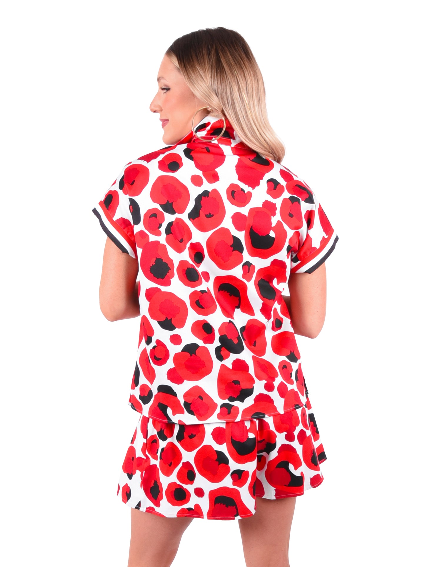 A person with long hair is facing away, wearing a matching short-sleeved shirt and skirt set featuring the Poppy Pullover’s bold red collegiate cheetah pattern on a white background, showcasing a tailored look against the plain white backdrop.