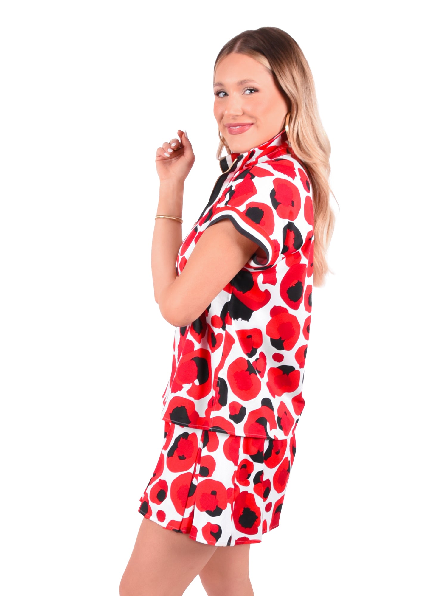 A woman with long blonde hair smiles while posing in a Poppy Pullover - Red Collegiate Cheetah featuring a 3/4 zip. Her matching shorts complete the tailored look as she stands against a white background.