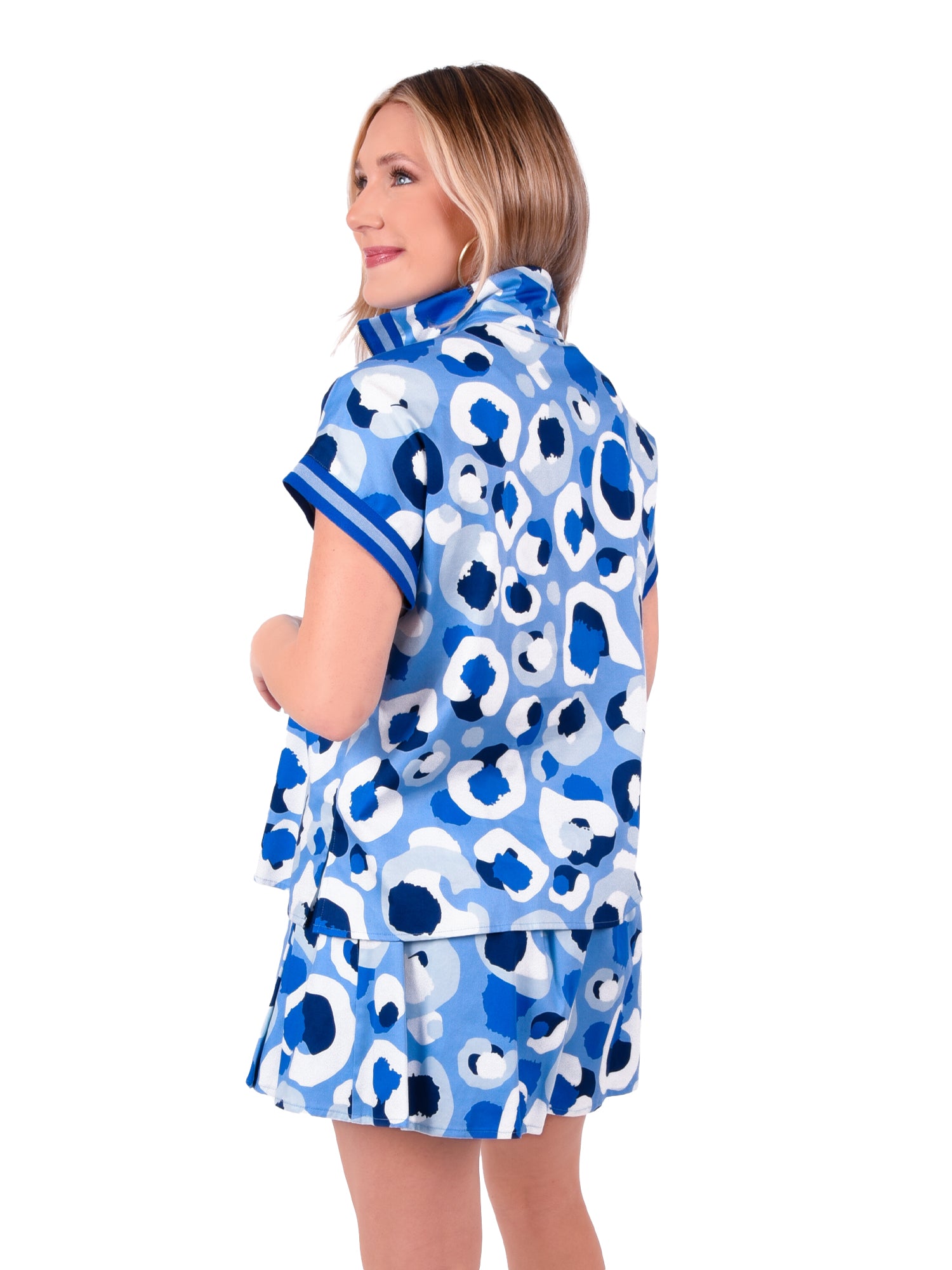 A woman with straight blonde hair wears a Poppy Pullover in the Blue Collegiate Cheetah pattern, paired with matching shorts, smiles while looking over her shoulder against a white background.