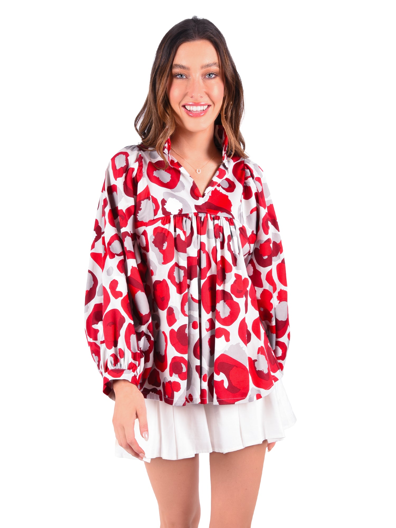 A person smiling, wearing a vibrant Stella Top in the Burgundy Collegiate Cheetah pattern with a ruffled collar. The long sleeves complement the white pleated skirt beautifully, making the outfit stand out against the white background.