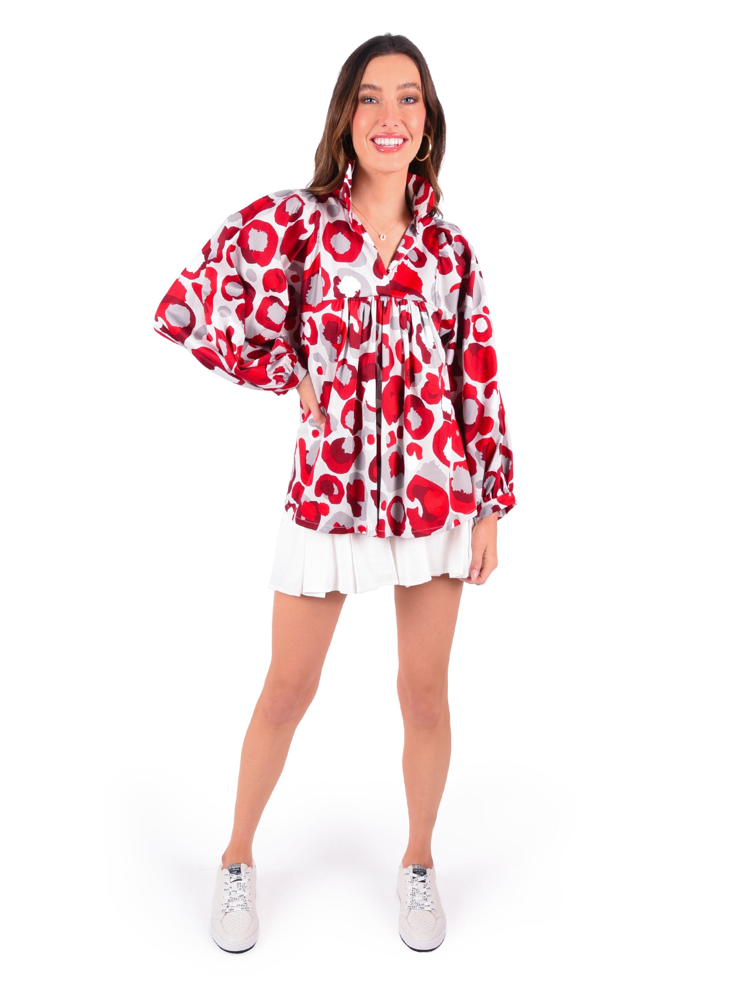 A woman is smiling and posing, wearing a stylish Stella Top in Burgundy Collegiate Cheetah featuring a ruffled collar, paired with a short white skirt. She has one hand on her hip and stands against a plain white background, complementing her look with chic white sneakers.