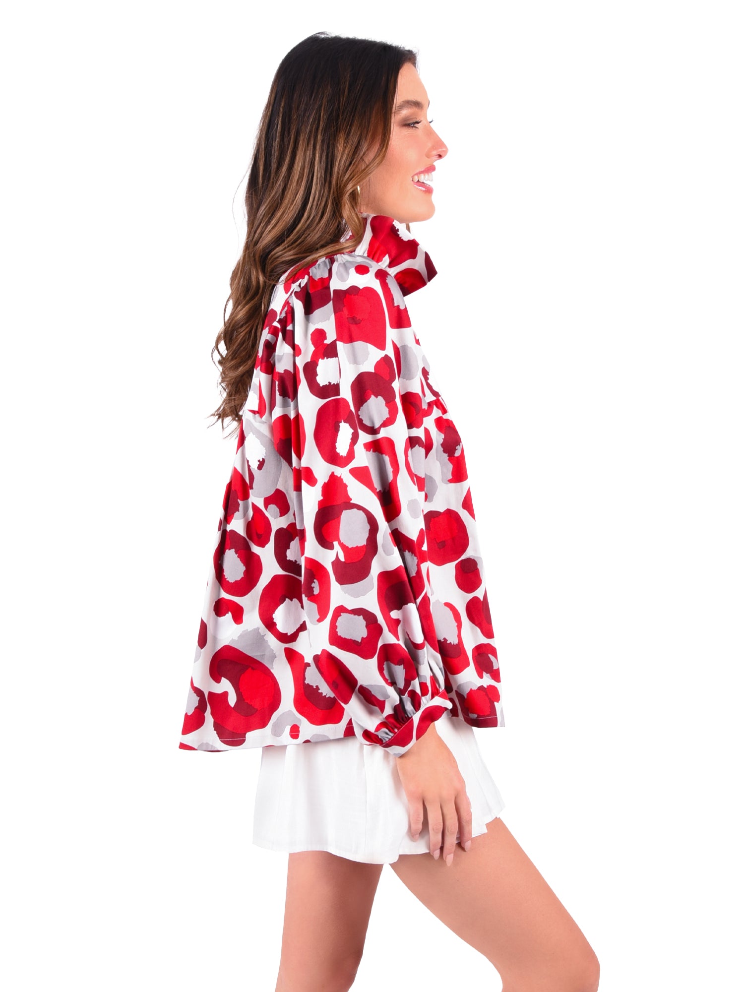 The woman is wearing the Stella Top in Burgundy with a Collegiate Cheetah pattern, a stylish long-sleeve blouse featuring a ruffled collar, paired with white shorts. She smiles warmly as her long hair cascades over her shoulders against the crisp white background.