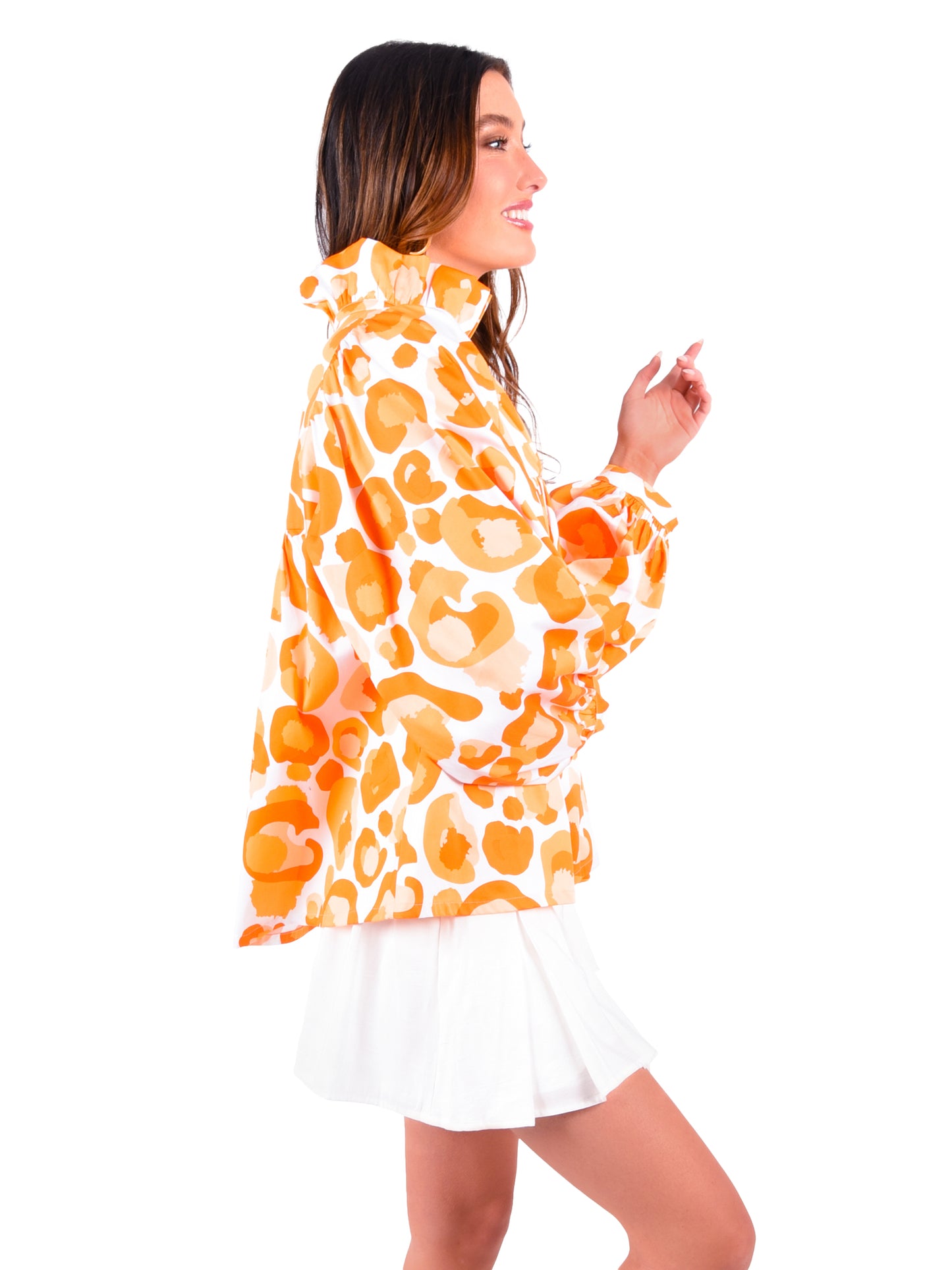 A woman in profile is wearing a Stella Top - Orange Collegiate Cheetah from the Emily McCarthy Collection, showcasing a relaxed empire shape with an orange and white cheetah pattern. The blouse features a ruffled collar and cuffs, and she has paired it with a white pleated skirt. She smiles and gestures with her hands against a plain white background.