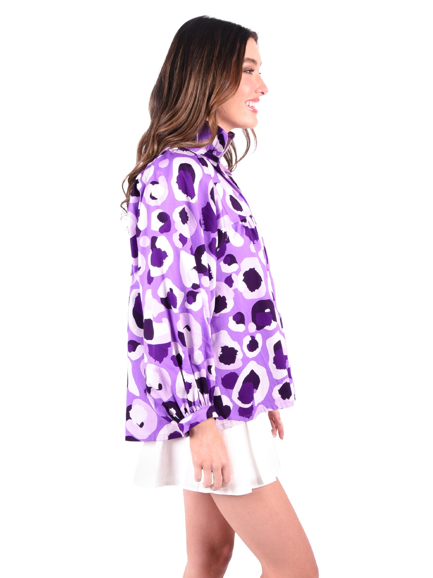 A woman stands smiling, wearing the Stella Top in Purple Collegiate Cheetah—an elegant long-sleeved shirt adorned with white and black abstract patterns. Her relaxed style is complemented by her long brown hair, as she pairs the top effortlessly with a white skirt. The plain white background accentuates her chic ensemble.