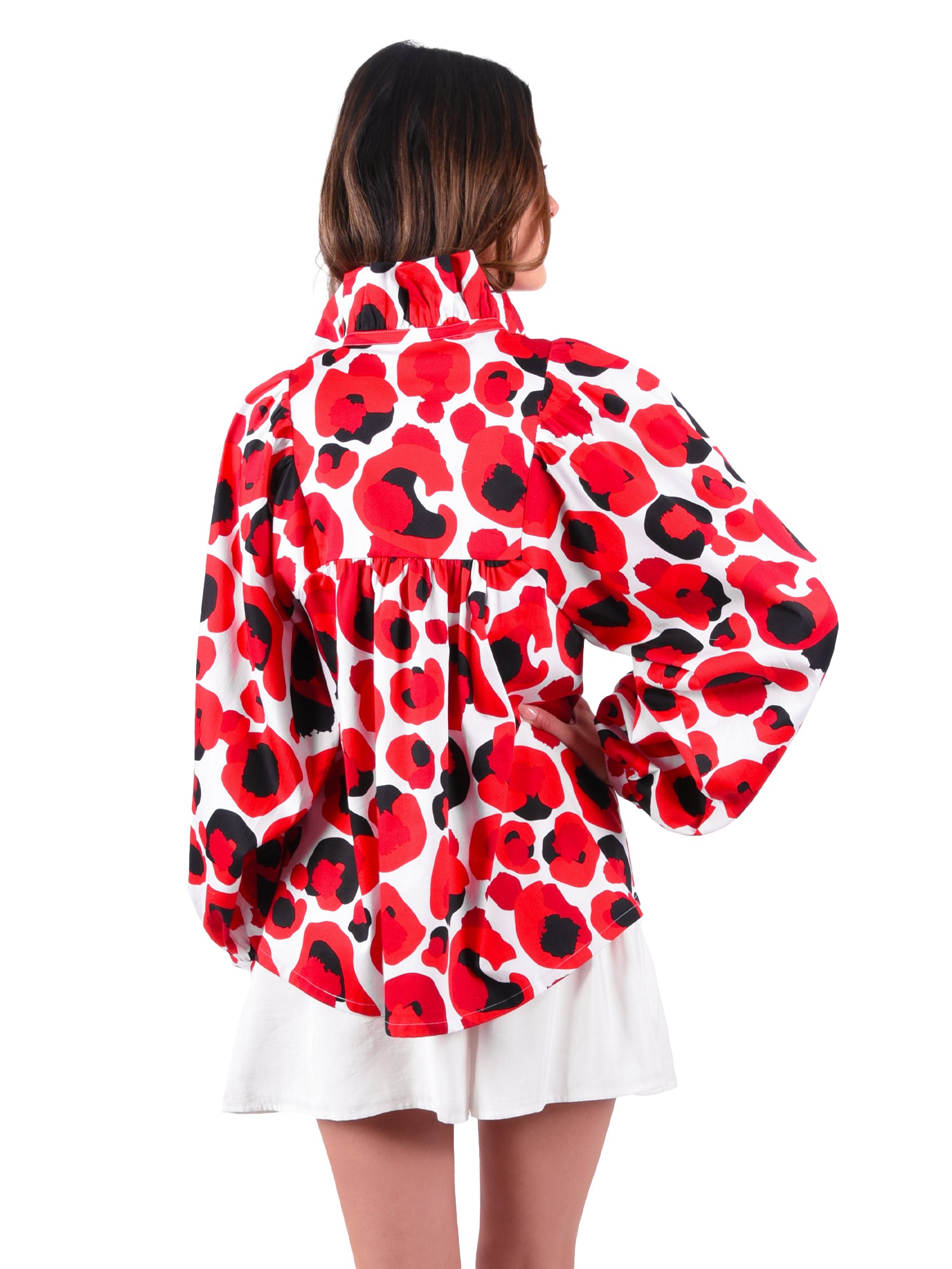 A woman is standing with her back to the camera, wearing the Stella Top in Red Collegiate Cheetah, which features a vibrant pattern with large floral designs and voluminous sleeves, paired with a white skirt. The signature loose fit enhances its relaxed empire shape.