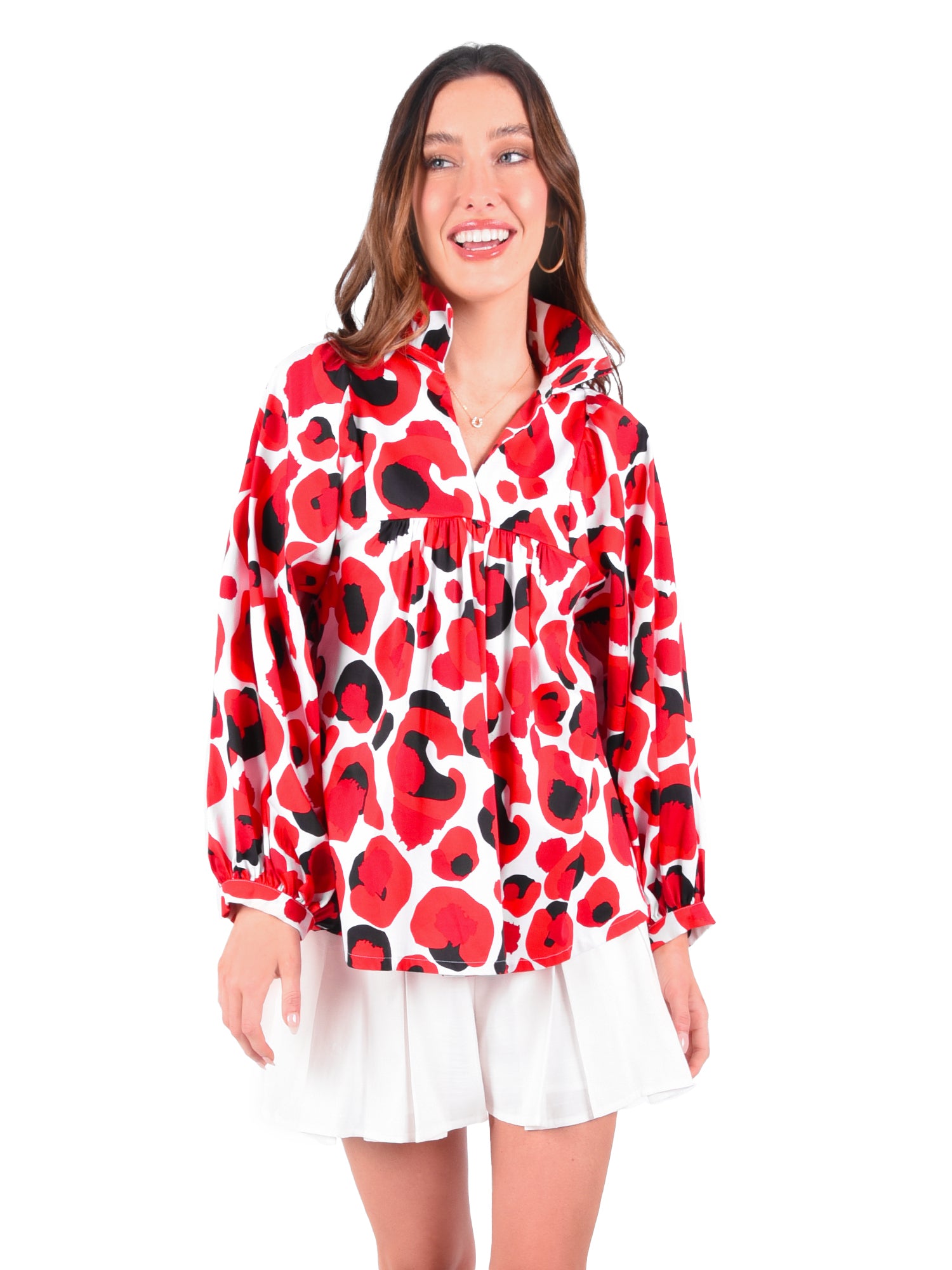 A person is smiling, wearing a vibrant Stella Top in the Red Collegiate Cheetah pattern, which features long sleeves and a relaxed empire shape. The top is paired with a white pleated skirt against a plain white background.