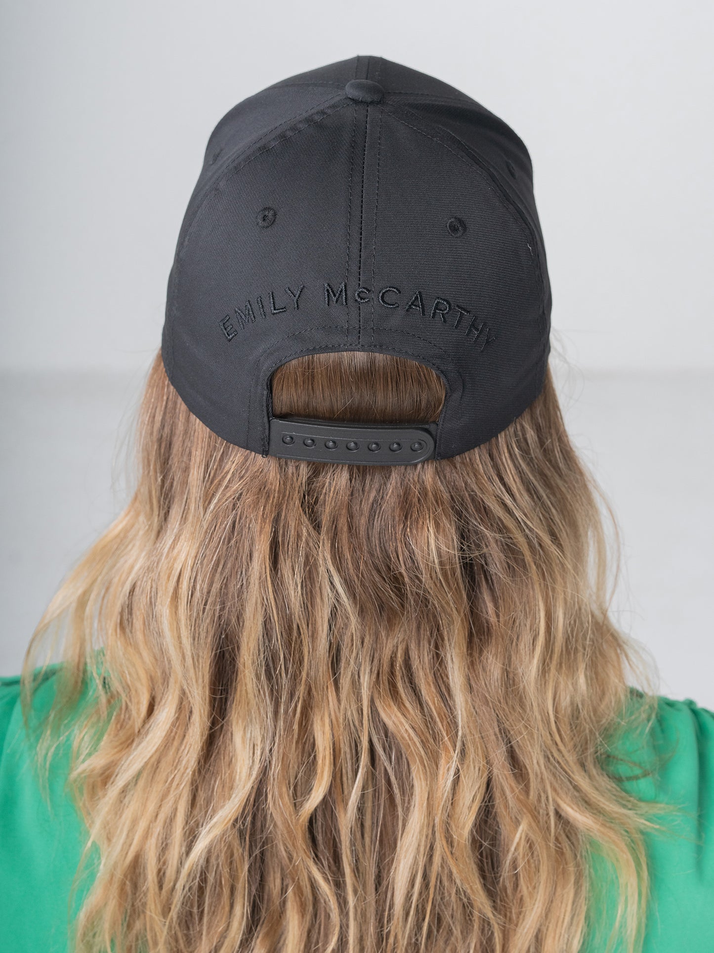 A person with long, wavy hair is seen sporting a black Emily McCarthy Signature Hat, featuring her name elegantly embroidered on the back. They pair this stylish cap with a comfortable green shirt while standing against a neutral gray background.